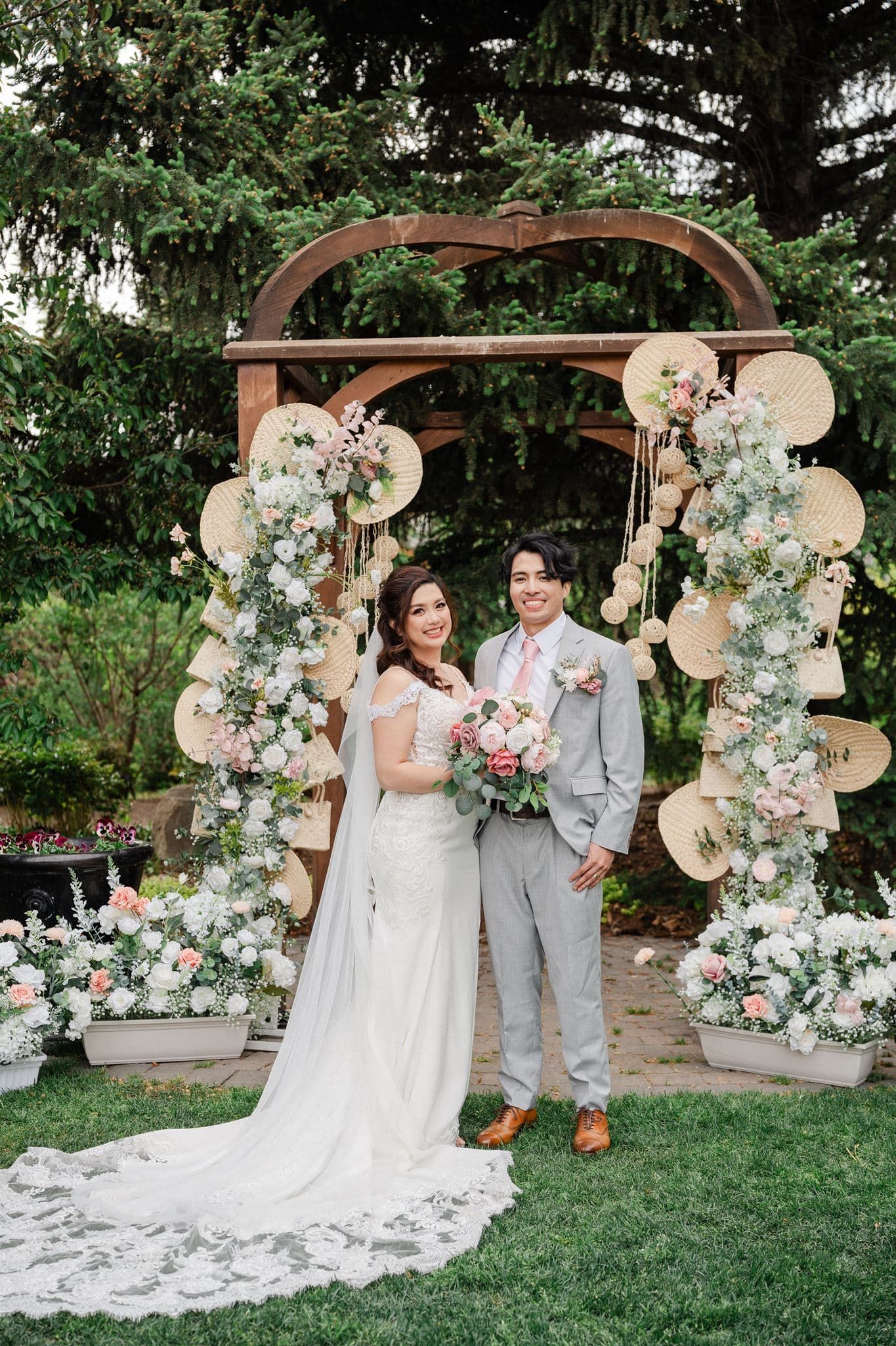 Bride and groom smiling under a floral arch with woven fans and pink blooms at an outdoor garden wedding ceremony.