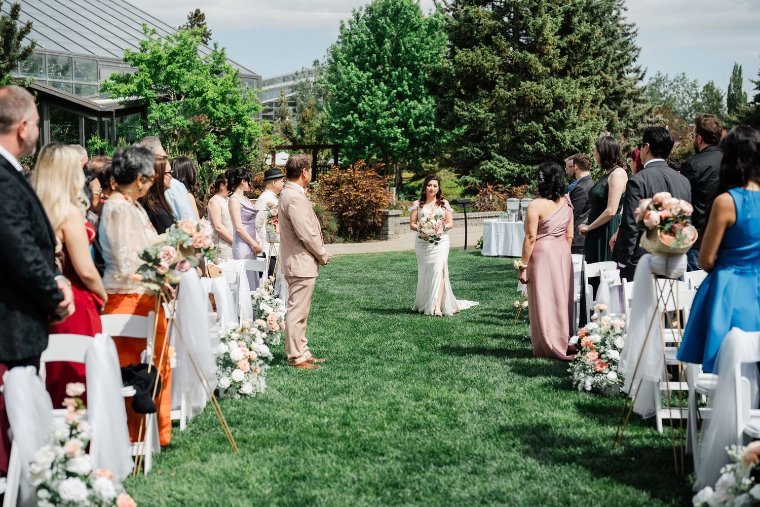 Outdoor wedding ceremony on green lawn with guests seated in white chairs, bridesmaids in pink dresses, and floral aisle decorations.
