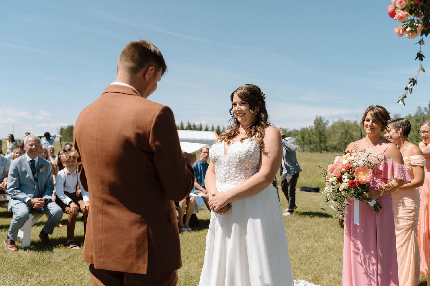 Bride and groom exchange vows at outdoor wedding ceremony with guests seated on sunny day.