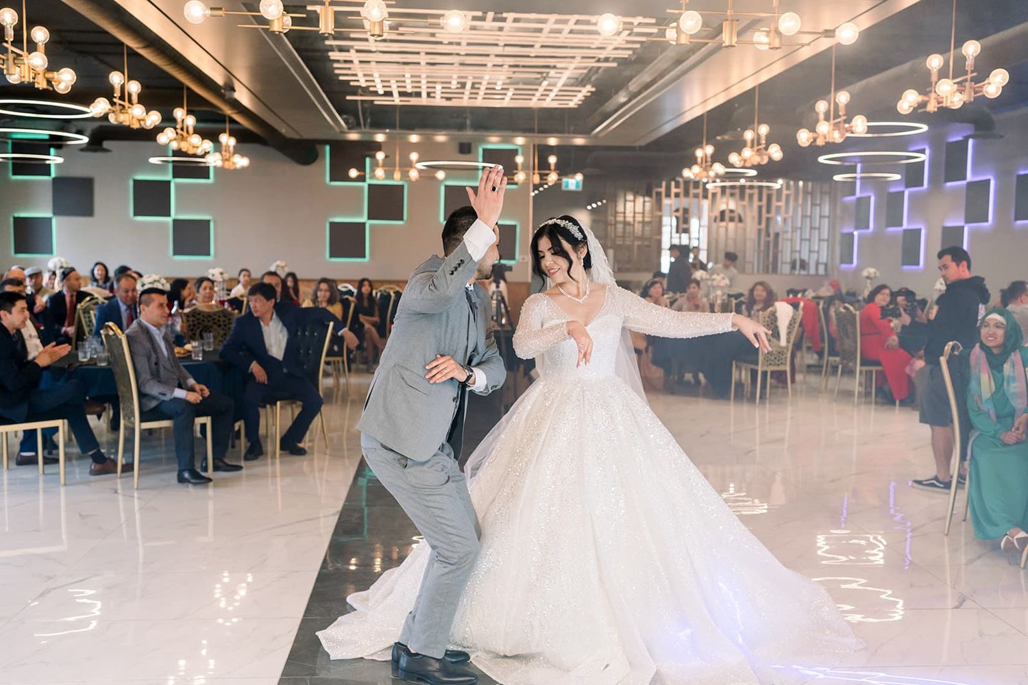 Bride and groom sharing first dance in elegant ballroom wedding reception with guests seated around dance floor.