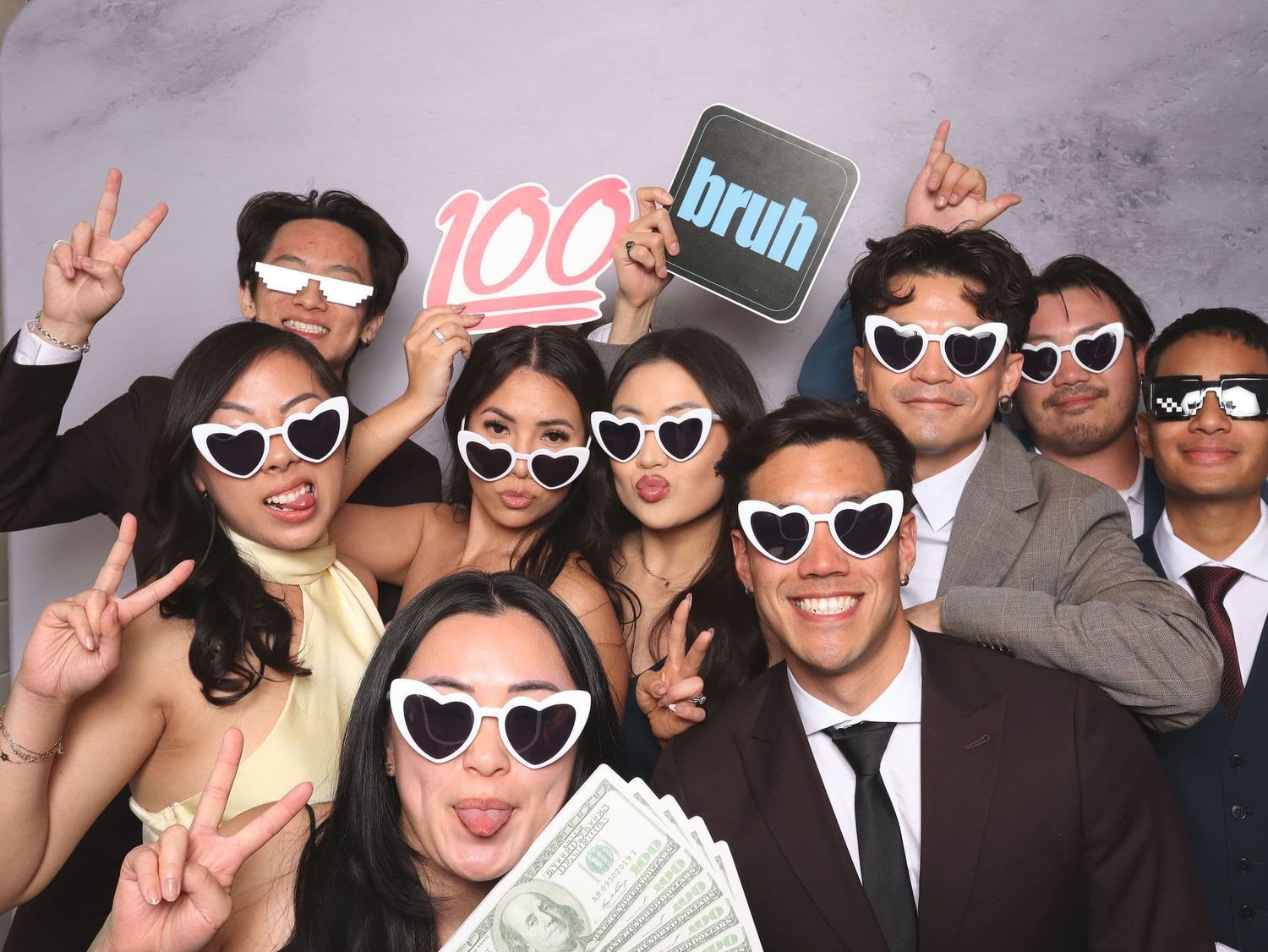 A fun group of friends pose with heart sunglasses, props, and cash in a photo booth.