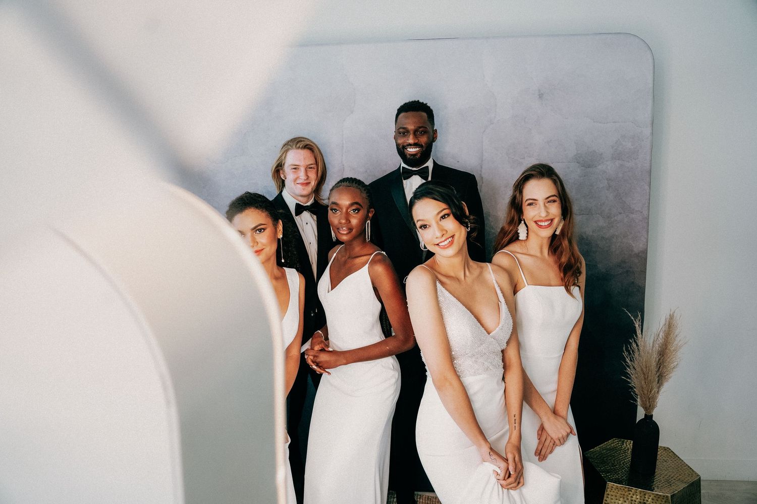 A group of wedding party members in black and white formal attire pose together in front of a gray backdrop for photos.