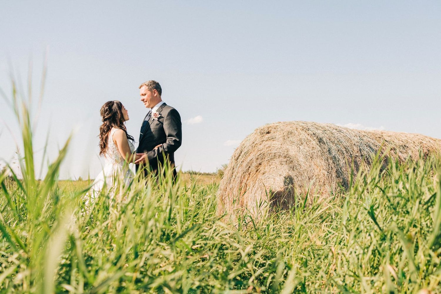 Bride and groom holding hands in a lush green field beside a rustic hay bale on a sunny wedding day.