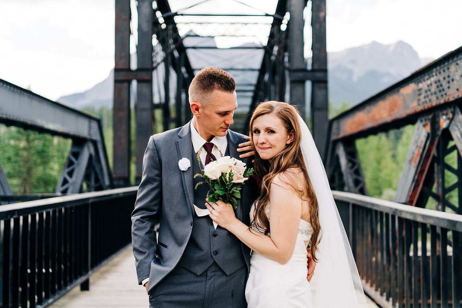 Bride and groom pose on a rustic iron bridge, she holds a white bouquet, mountains visible in background.
