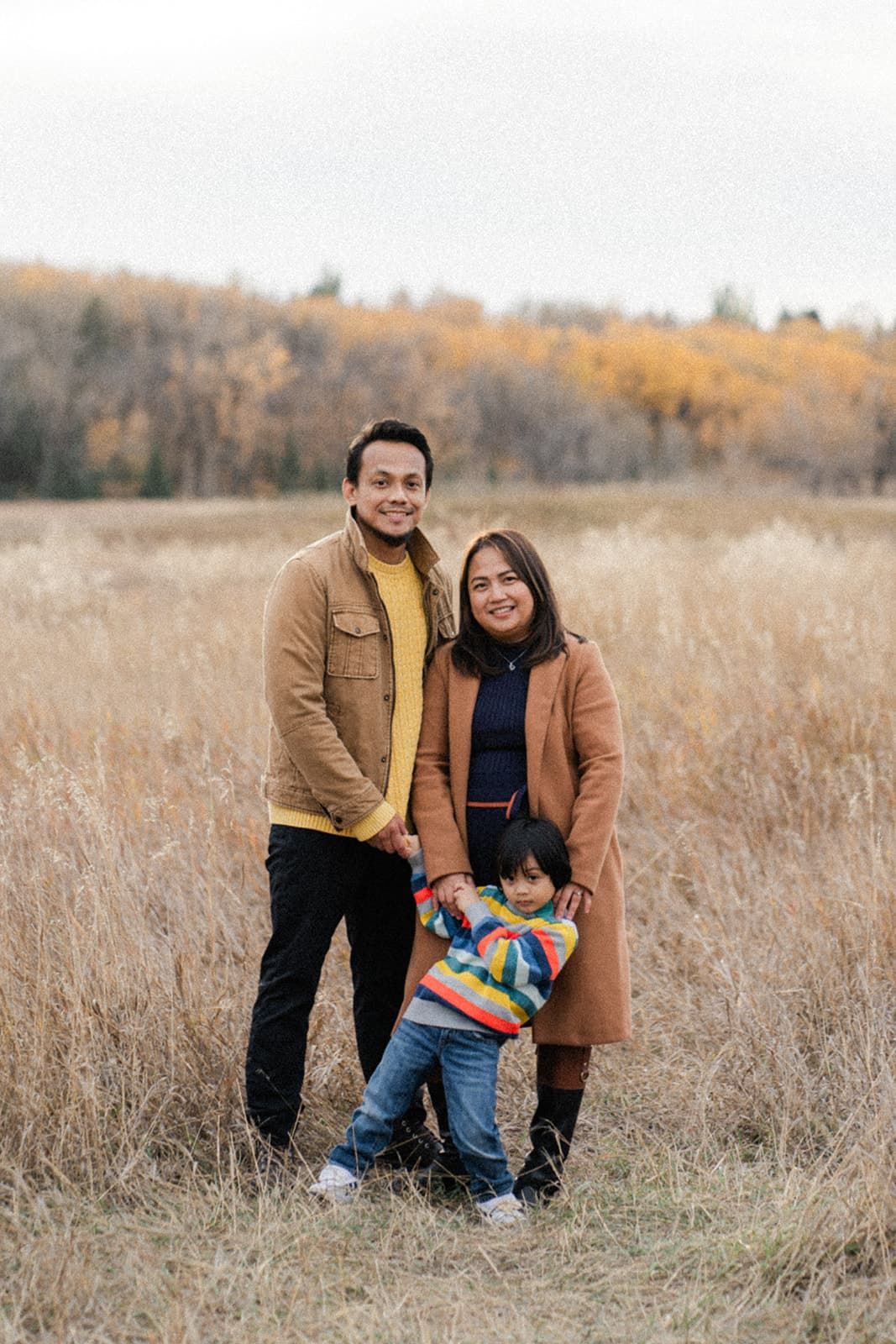 Family of three posing in an autumn field with golden trees, parents in brown coats with young child in colorful striped jacket.