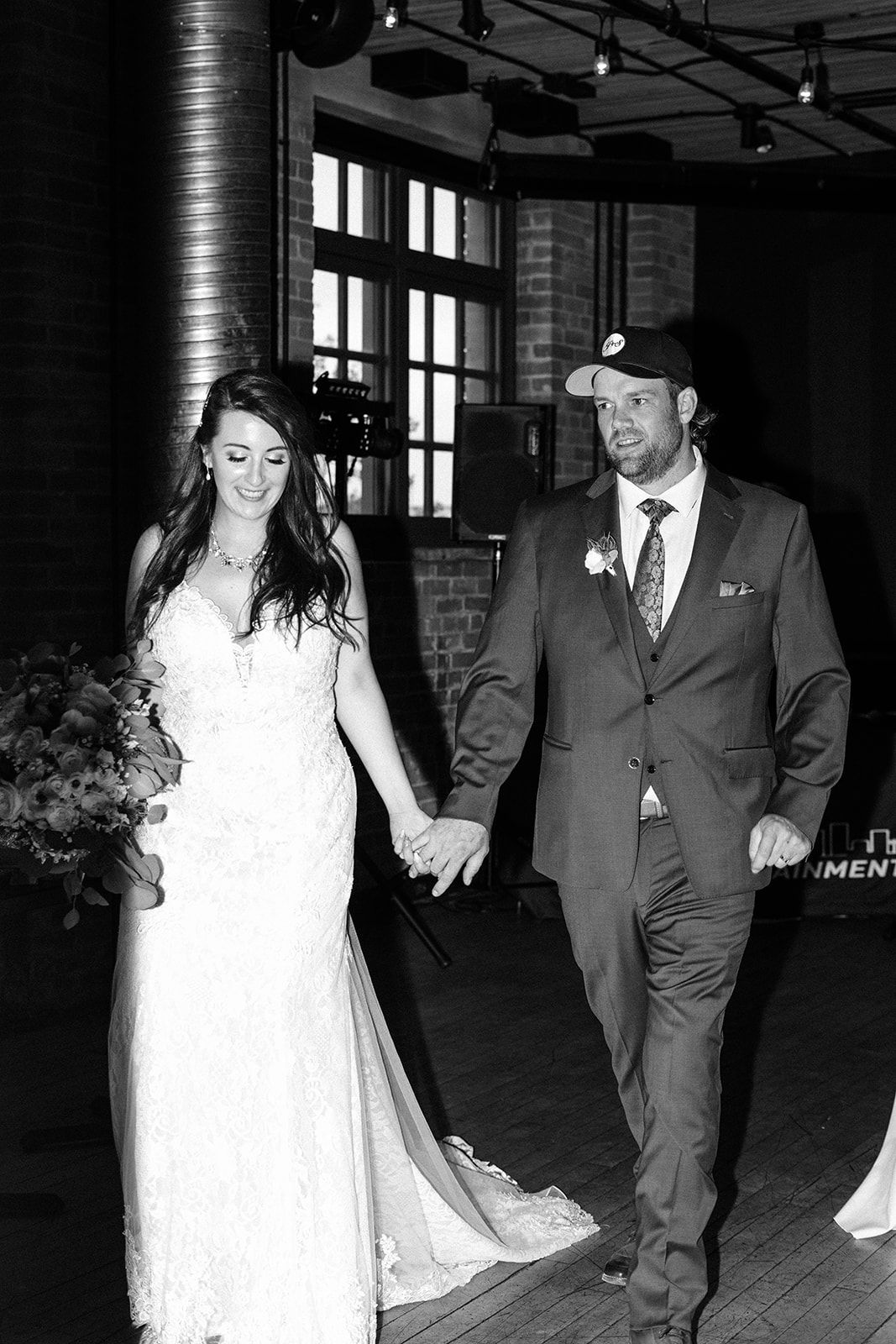 Black and white photo of a bride in lace gown and groom in suit holding hands walking at their wedding reception.
