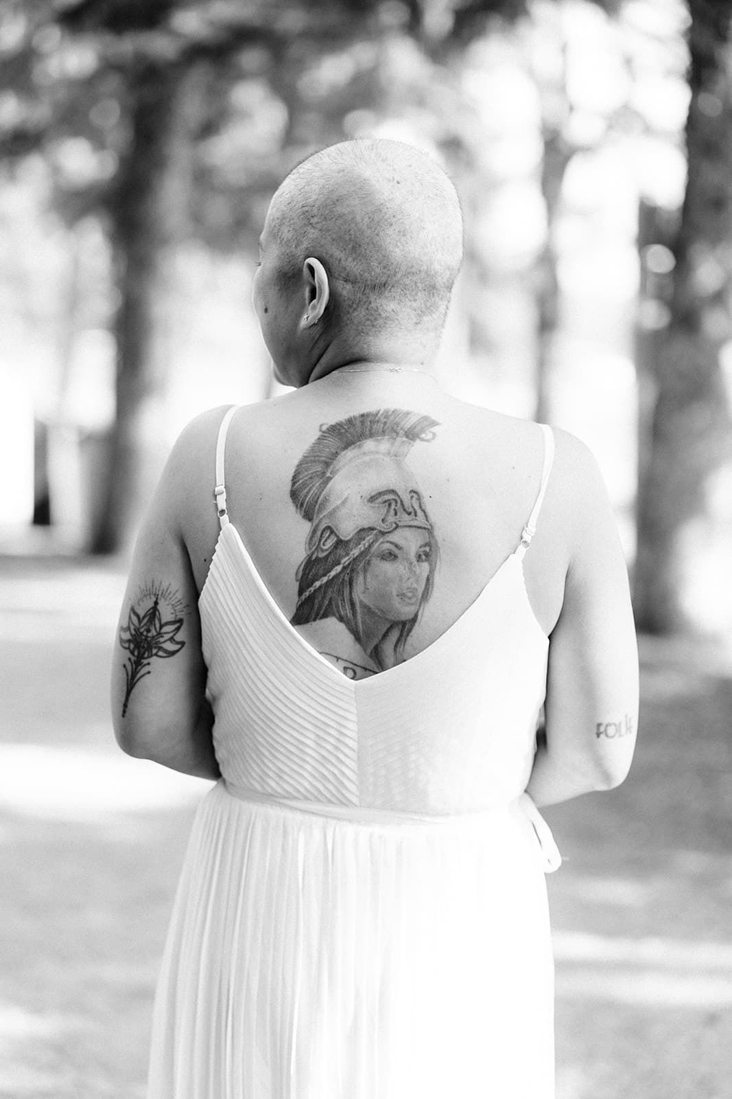 Black and white photo of a bald woman in white dress showing back tattoo of a Roman warrior goddess outdoors.