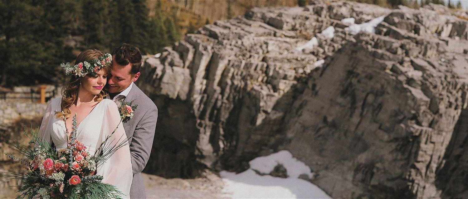 A romantic couple embraces near rocky cliffs at sunset with mountains and natural scenery in the background.