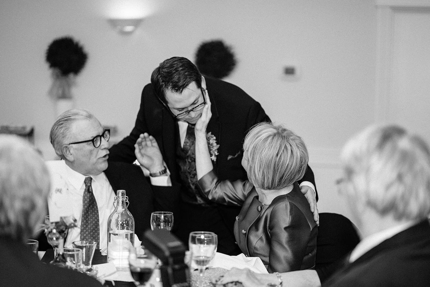 Groom greets seated guests at wedding reception in elegant black and white candid photography.