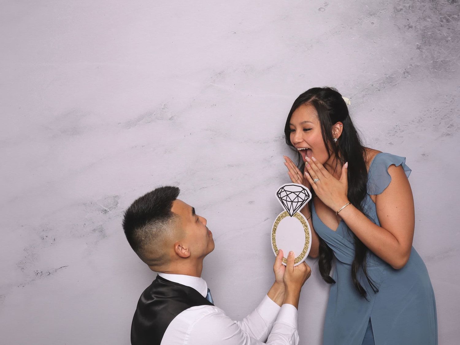 Man on one knee holding a ring prop while woman reacts with surprise in a romantic photo booth moment.