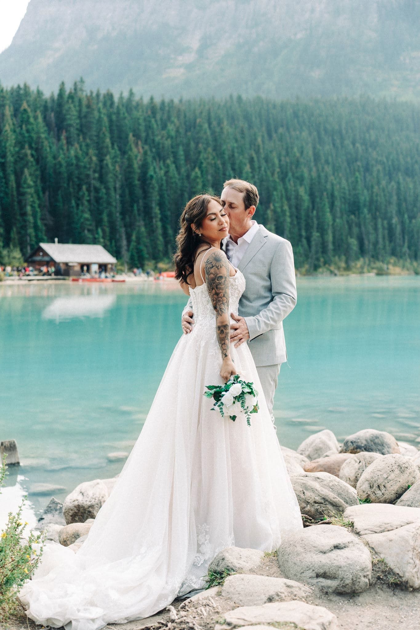 Bride and groom embrace on rocky lakeside with turquoise water, pine forest, and mountain backdrop.