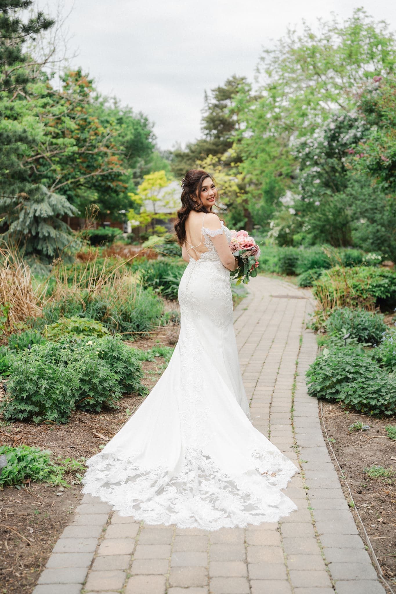 Bride in a lace mermaid wedding gown with a train poses on a garden path holding a pink bouquet.