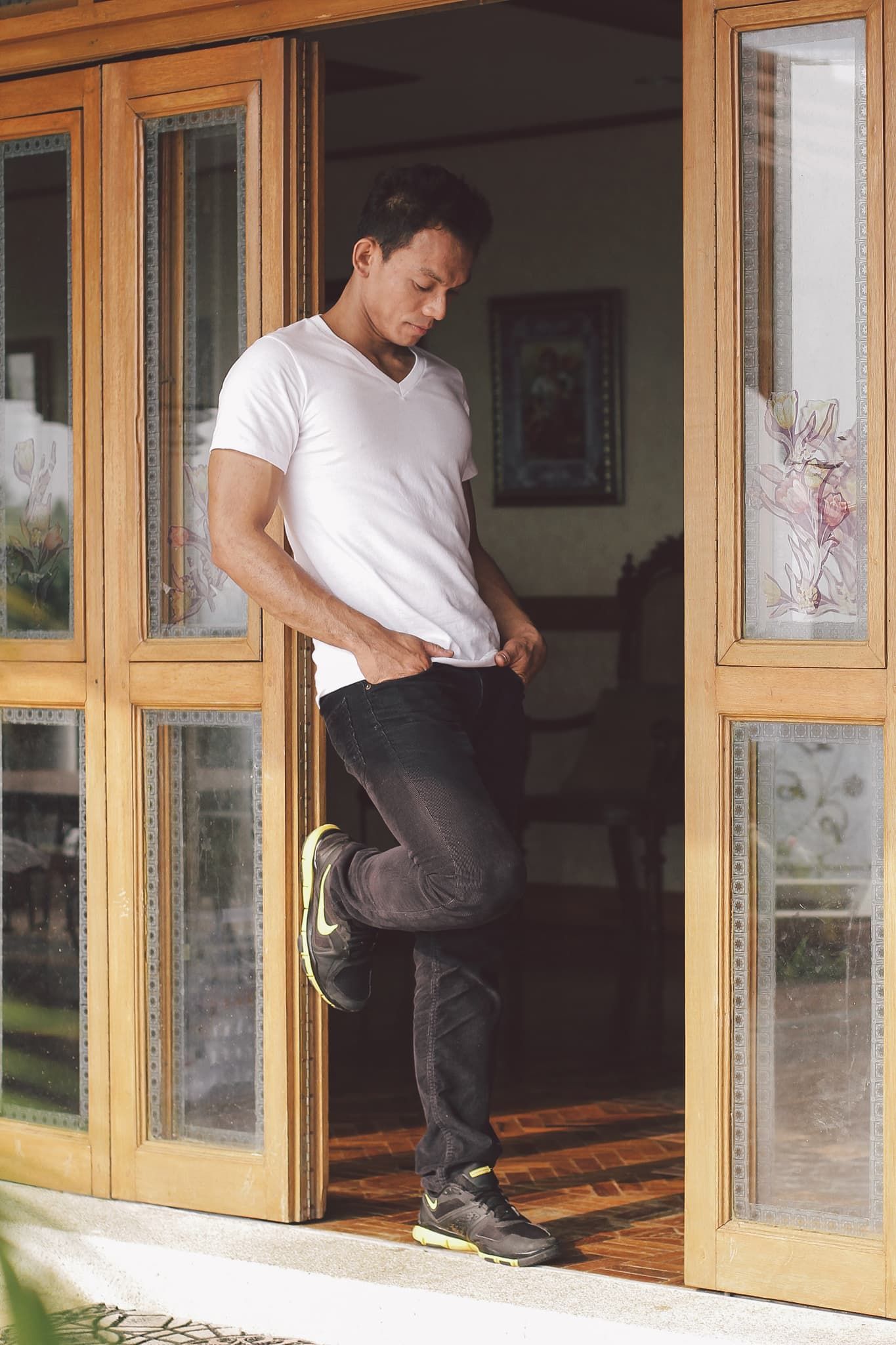 Man in white t-shirt and black jeans standing in wooden glass doorway, looking down while adjusting shirt.