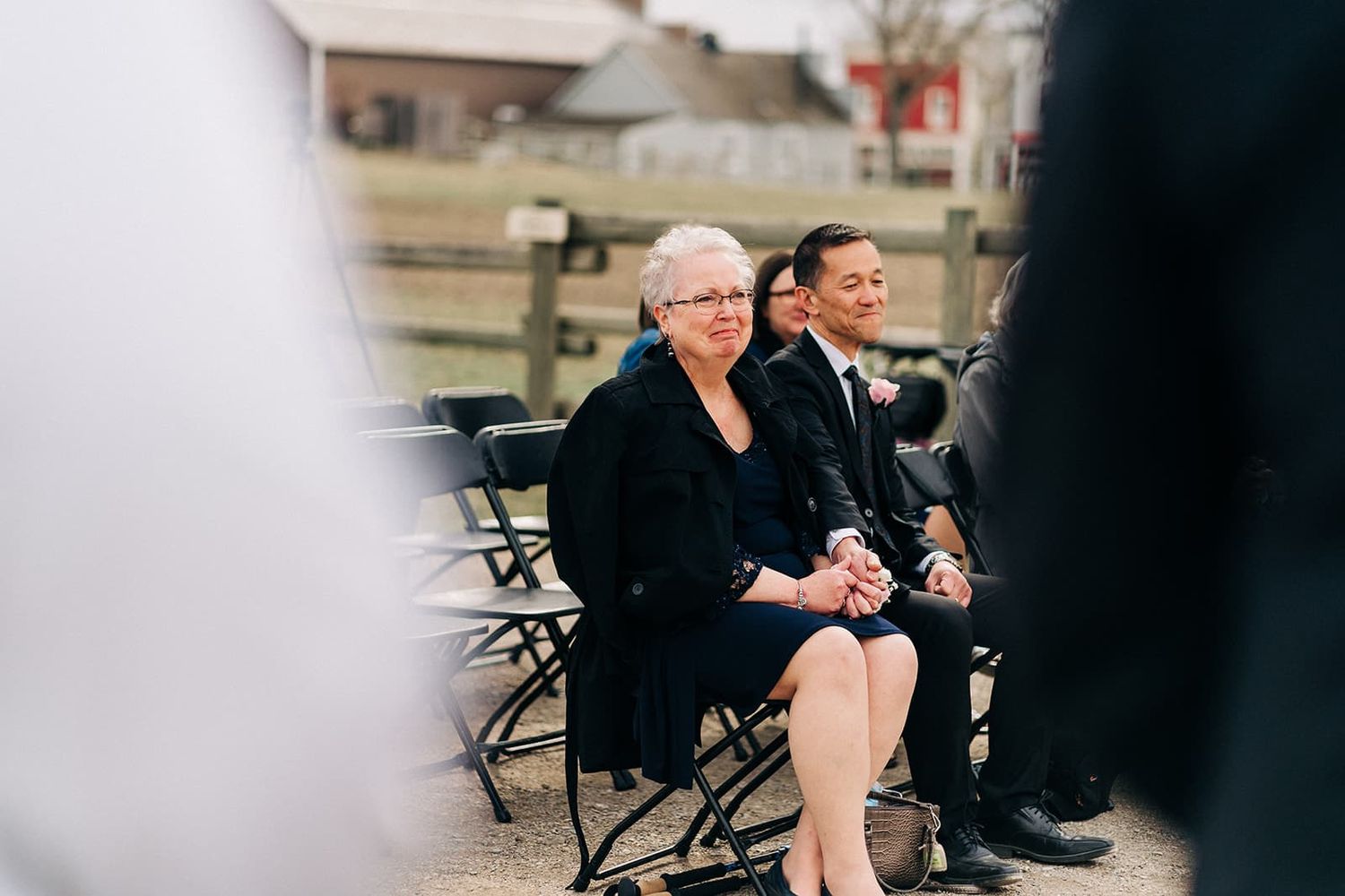 Wedding guests seated in chairs watch an outdoor wedding ceremony at a rustic venue.