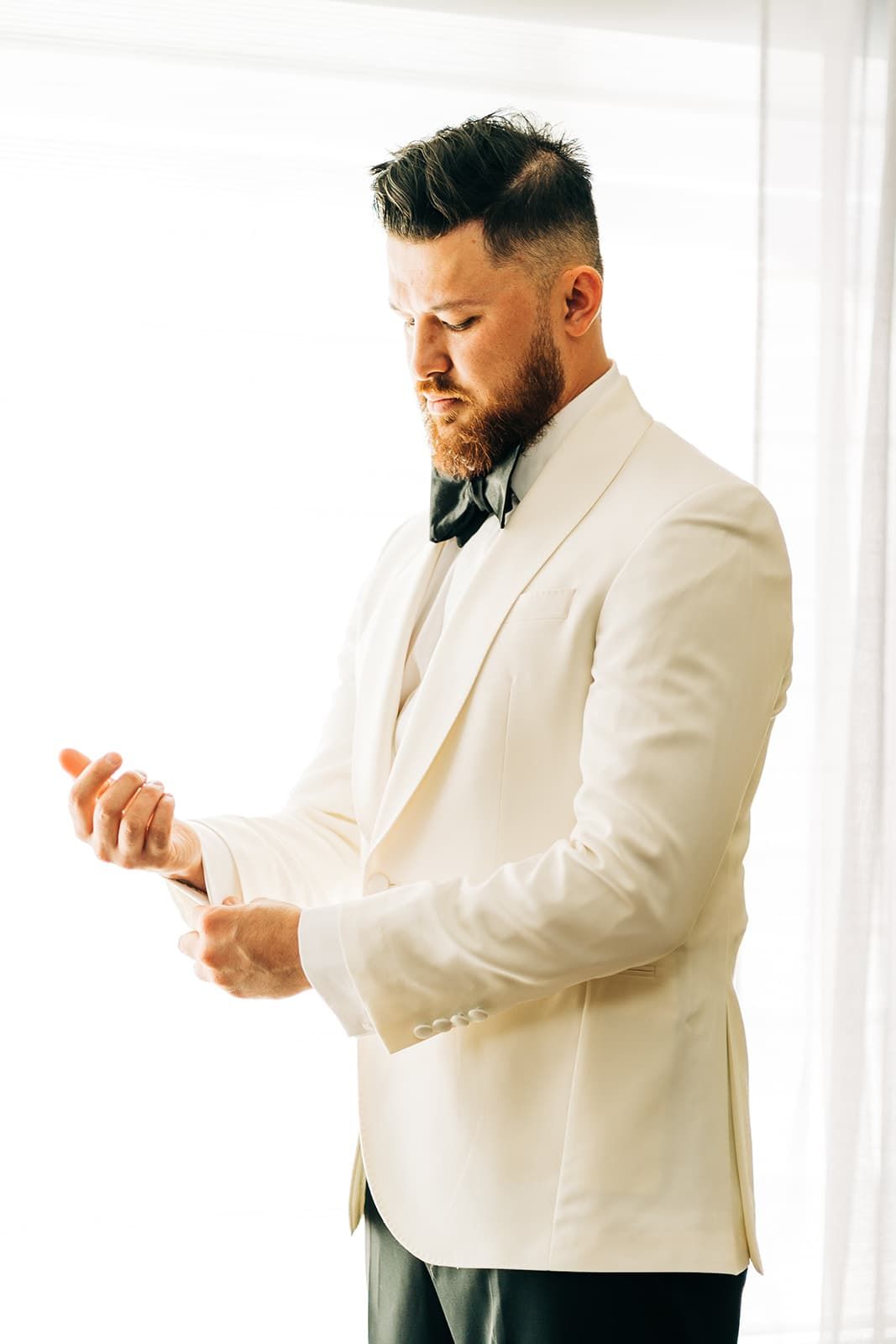 Elegant groom in white tuxedo jacket and black bow tie adjusting his cufflinks before the wedding ceremony.