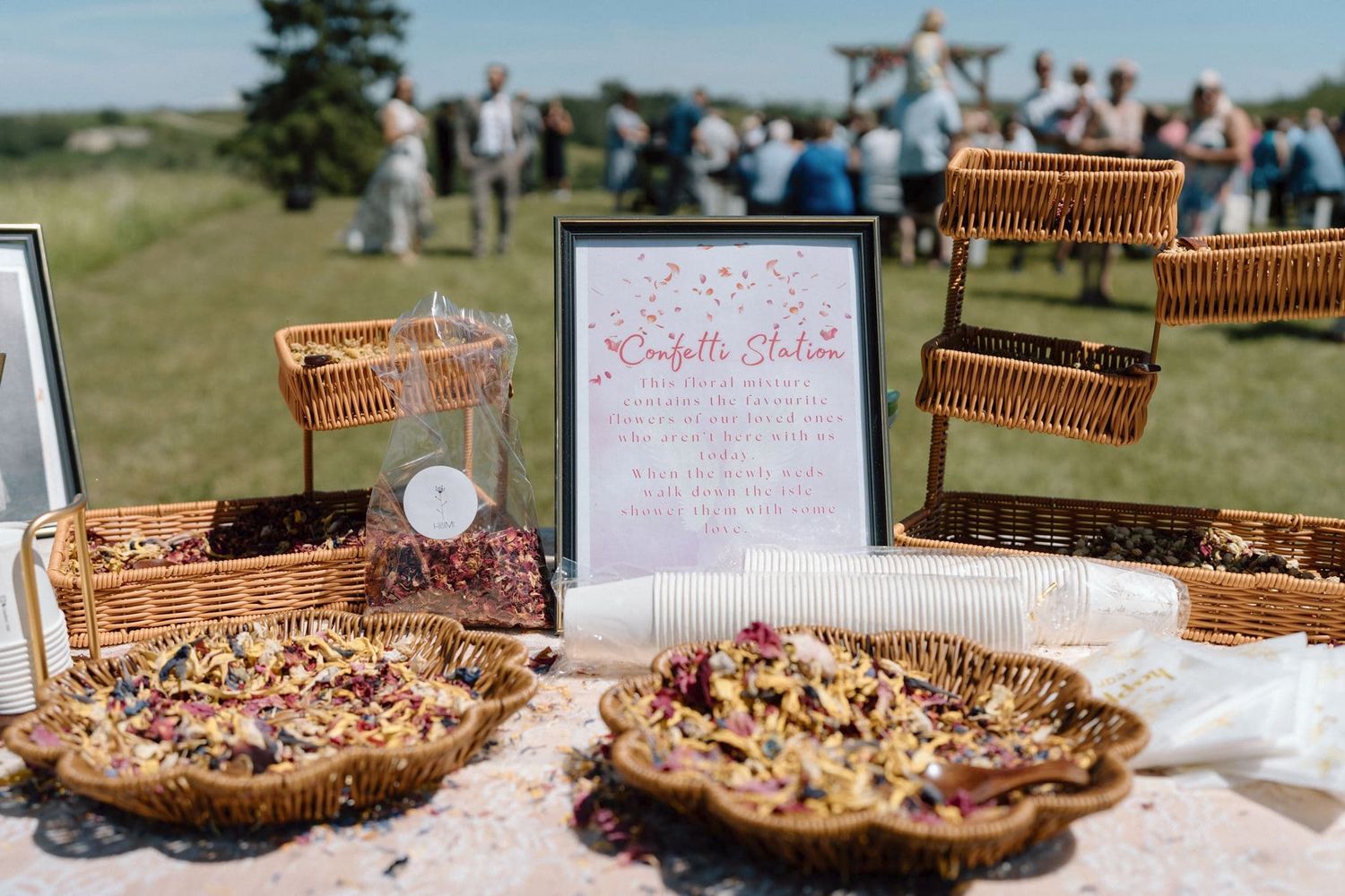 Confetti station with wicker baskets and bowls of flower petals at an outdoor wedding reception.