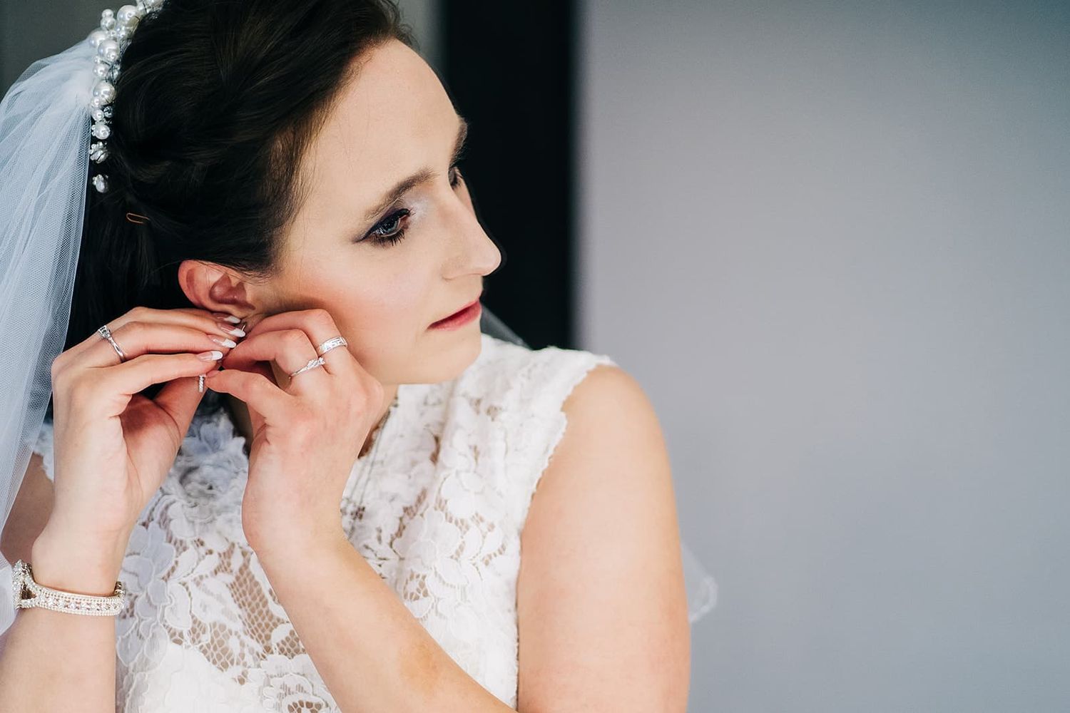 Elegant bride in lace dress adjusts pearl earring while preparing for her wedding ceremony.