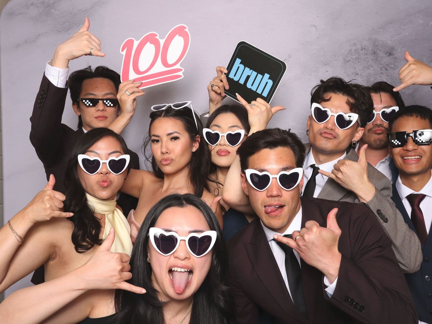 Group of friends posing at wedding photo booth with fun props including heart sunglasses and emoji signs.