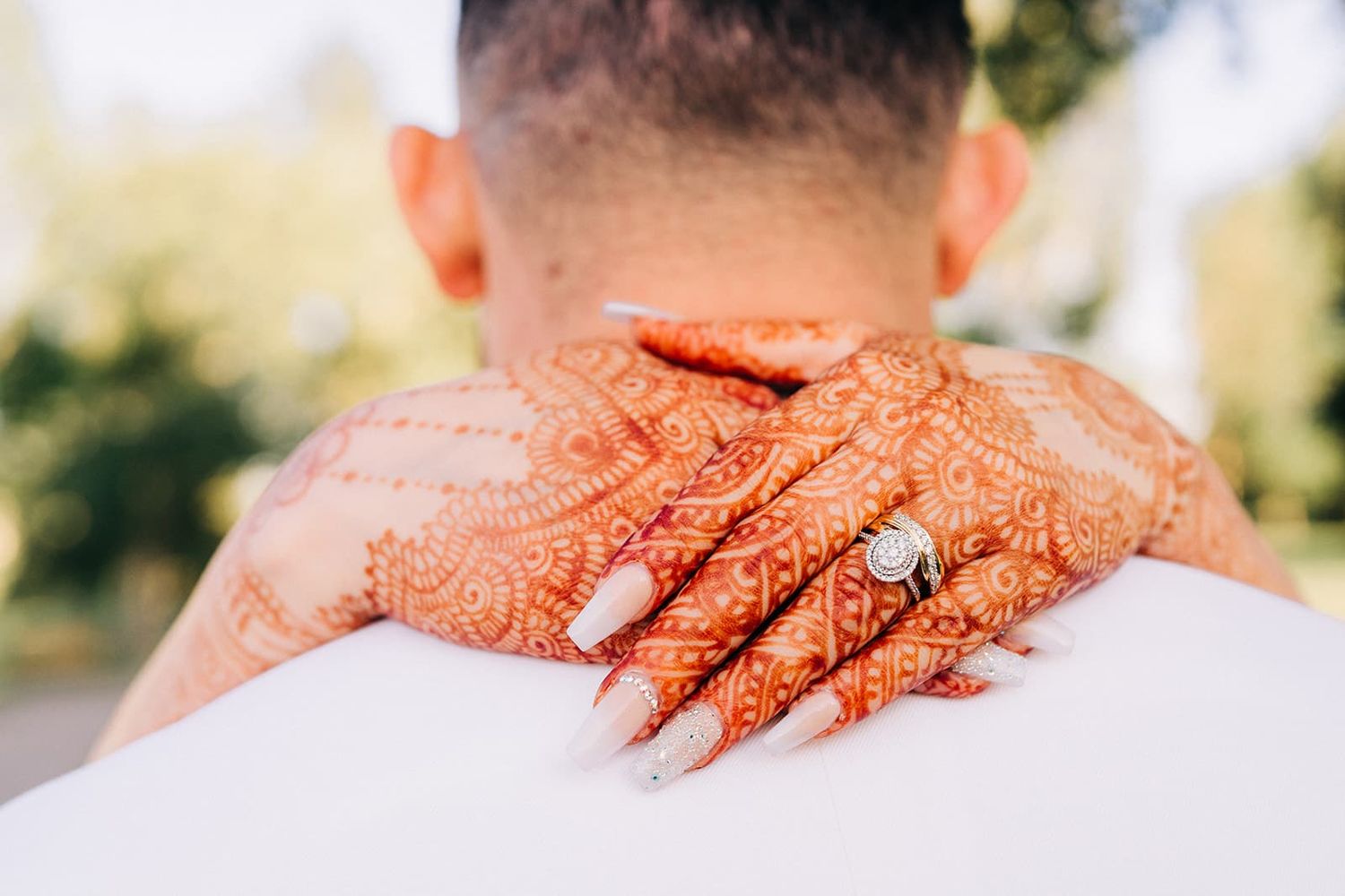 Bride's henna-adorned hands with diamond engagement ring resting on groom's shoulders at outdoor wedding.