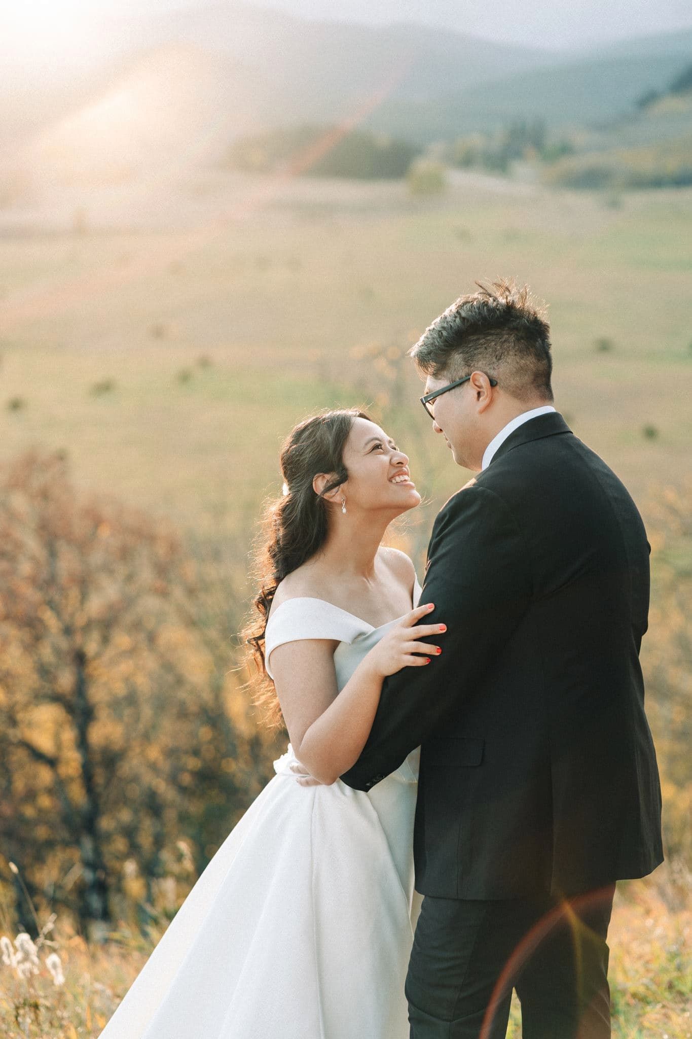 A bride in a white off-shoulder gown and groom in a black suit embrace at golden hour with scenic mountains behind.