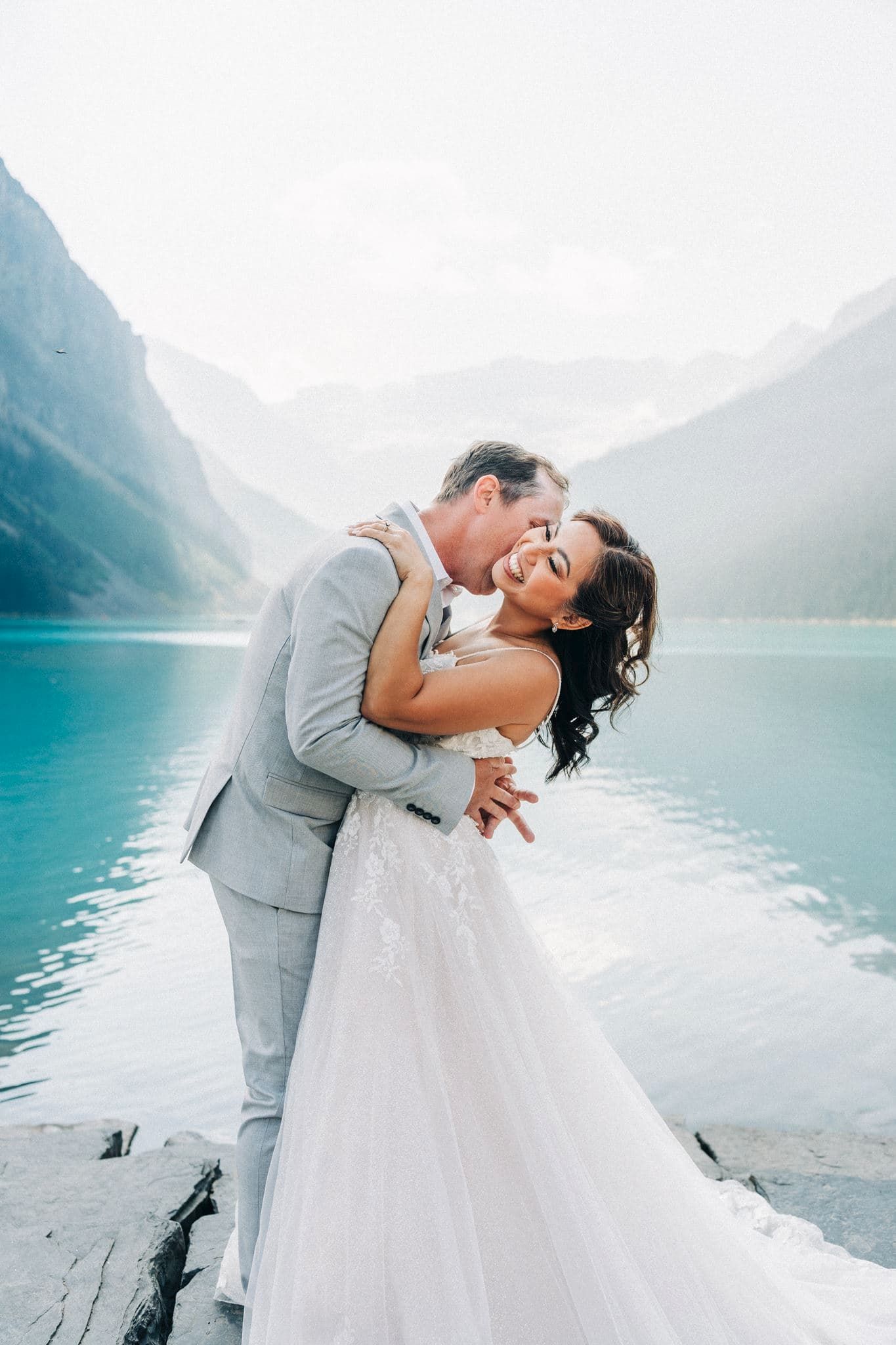 Bride and groom embrace romantically by turquoise Lake Louise with misty mountains in background on their wedding day.