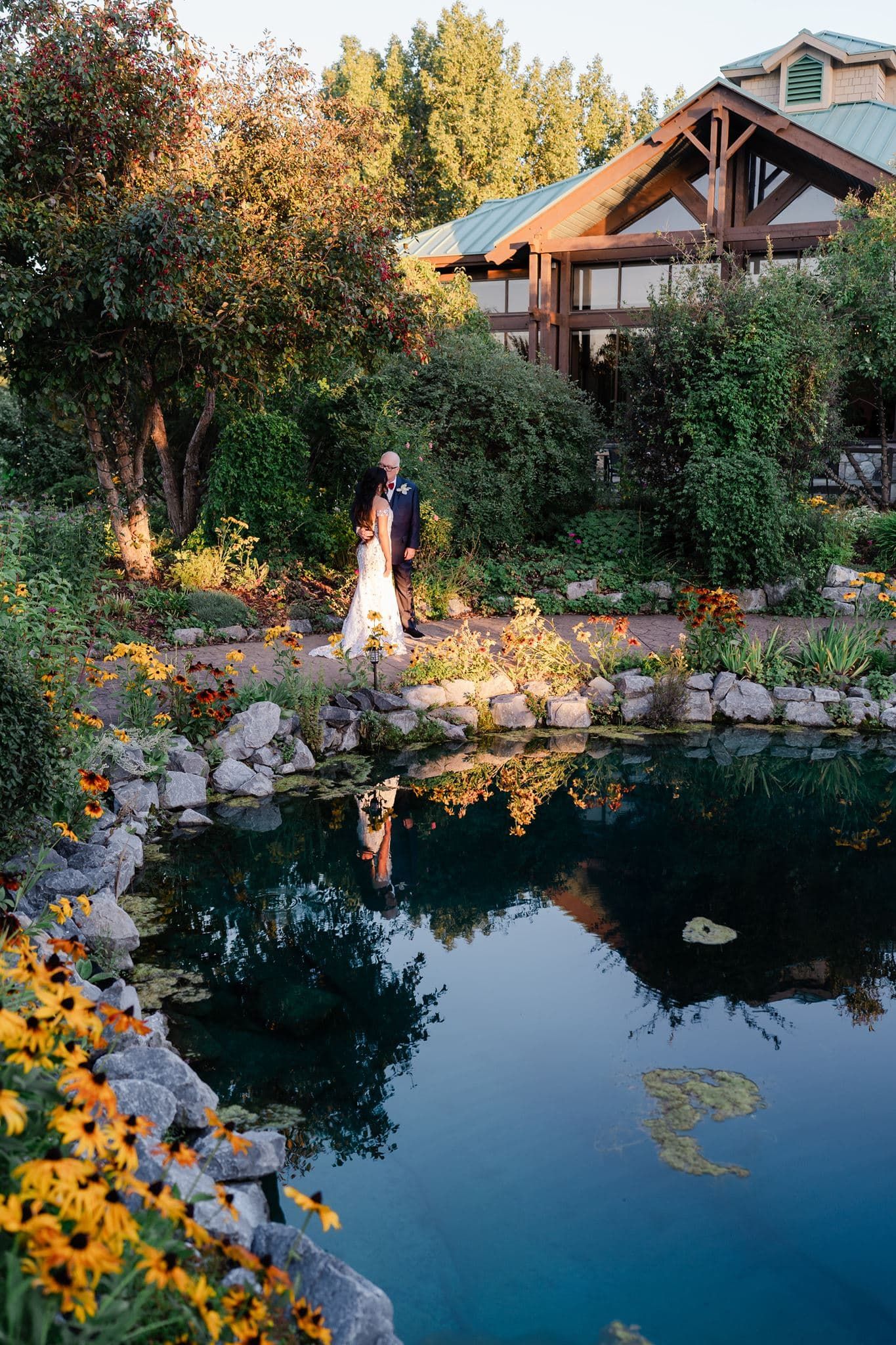 Bride and groom embrace beside a rock-lined reflective pond with colorful flowers and a rustic timber lodge in the background.