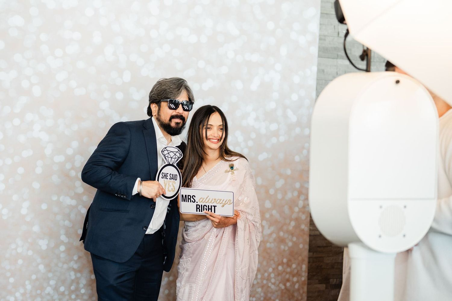 Couple posing at wedding photo booth holding Mr. and Mrs. signs, man in navy suit, woman in pink saree.