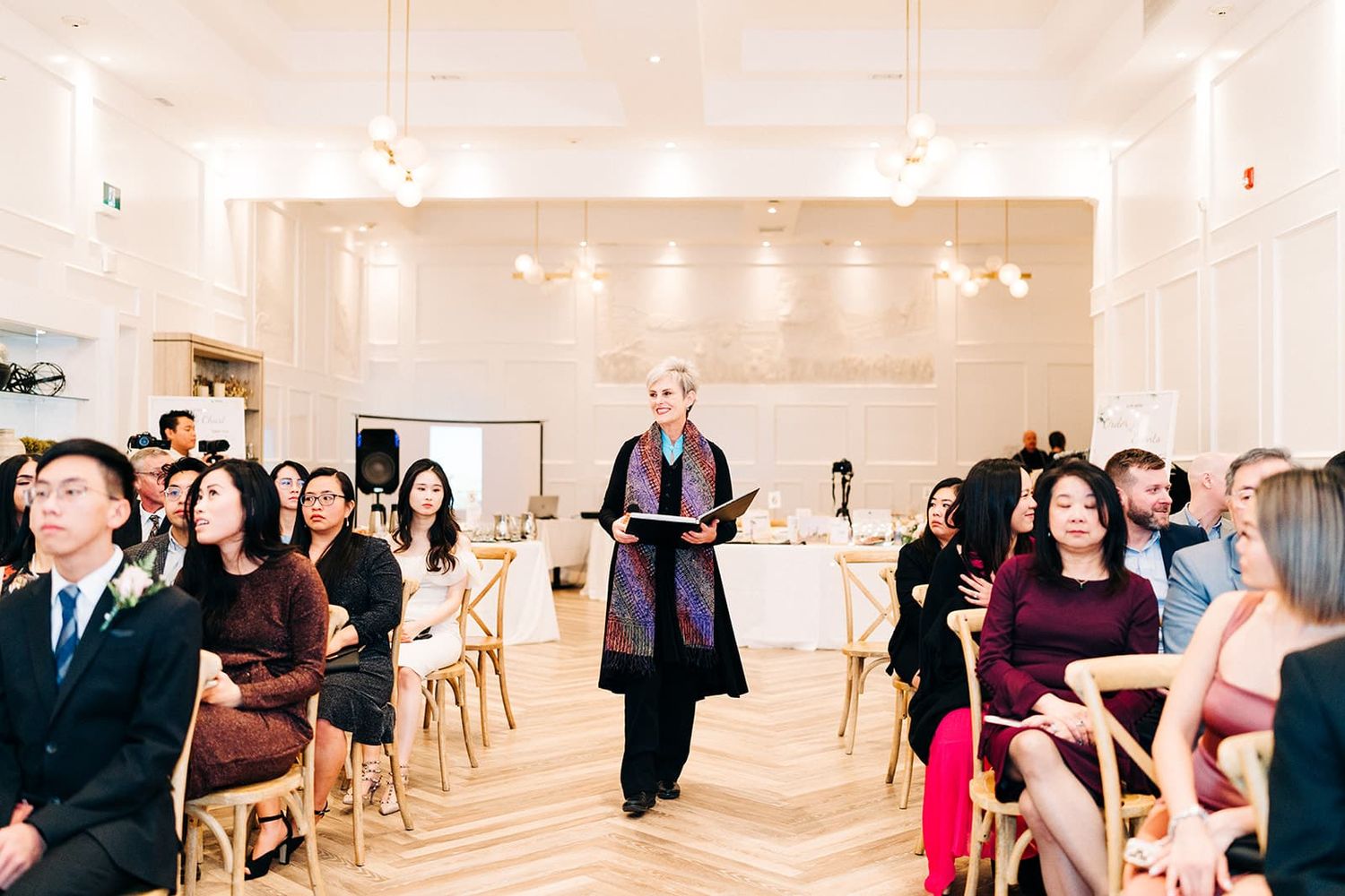Officiant walks down aisle at elegant indoor wedding ceremony with seated guests in a bright white venue.