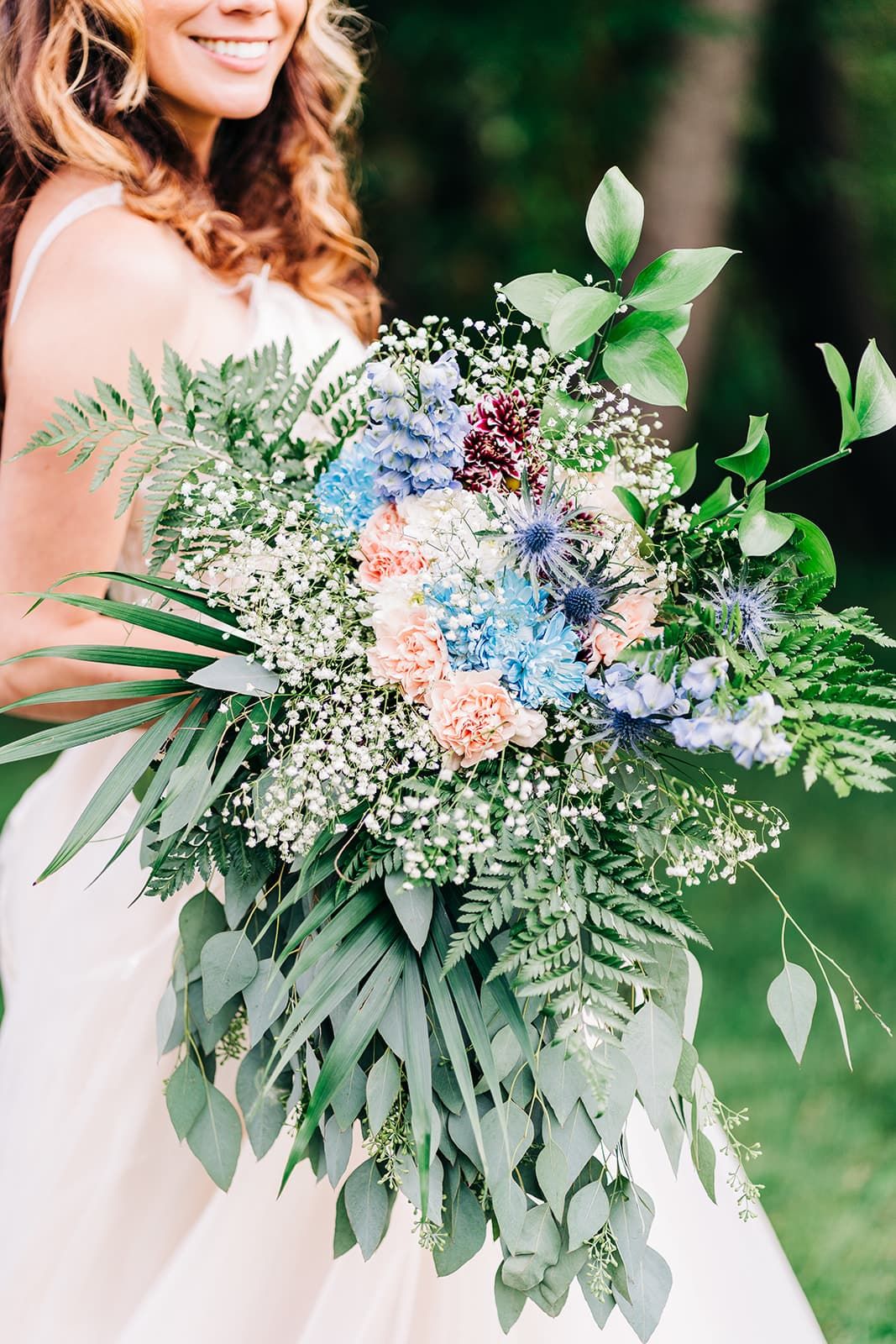 Smiling bride holding a lush wedding bouquet with blue, peach flowers, ferns, and baby's breath outdoors.