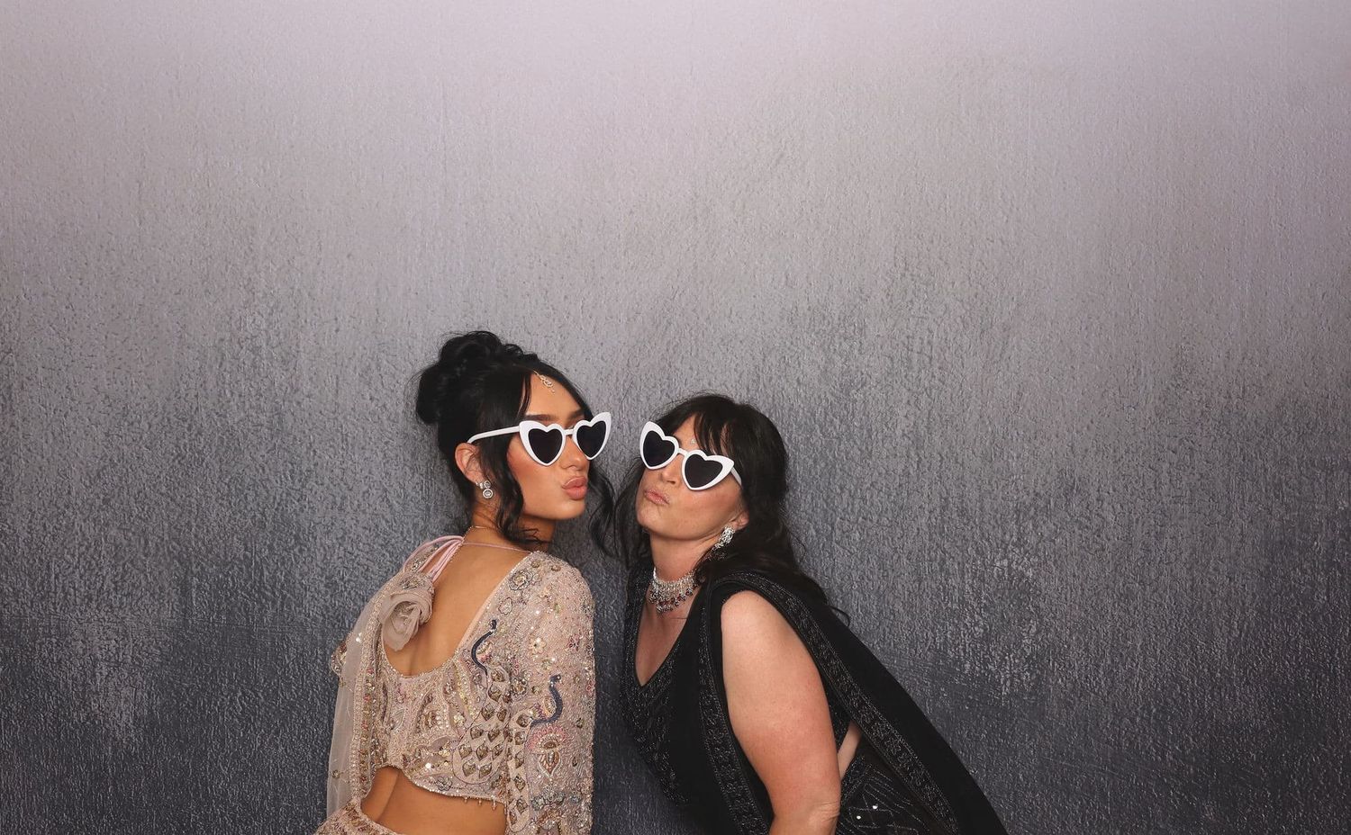 Two women wearing white heart-shaped sunglasses pose together against a gray backdrop.