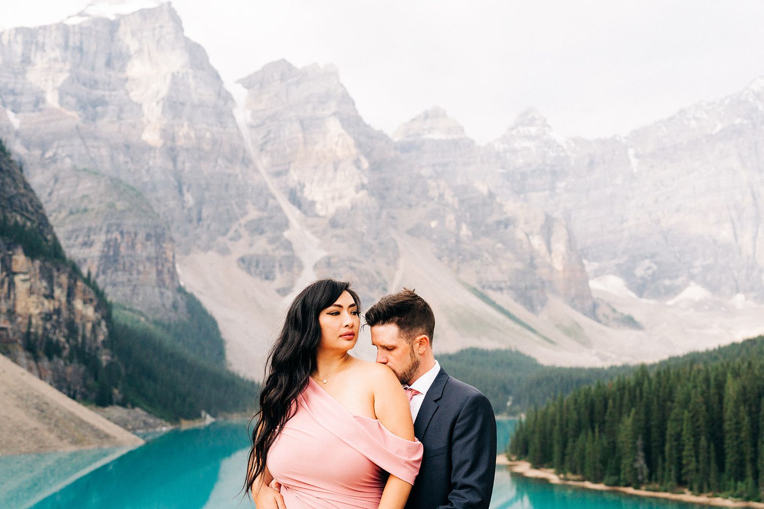 A romantic couple embraces in front of turquoise Lake Moraine with majestic Rocky Mountains in the background.