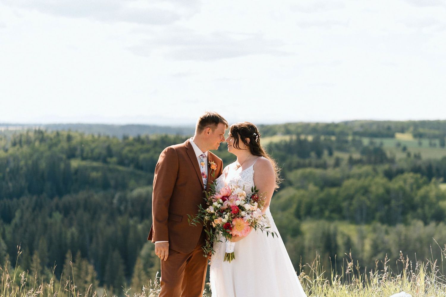 Bride and groom share a romantic moment on a hilltop with lush green forest and cloudy sky backdrop.