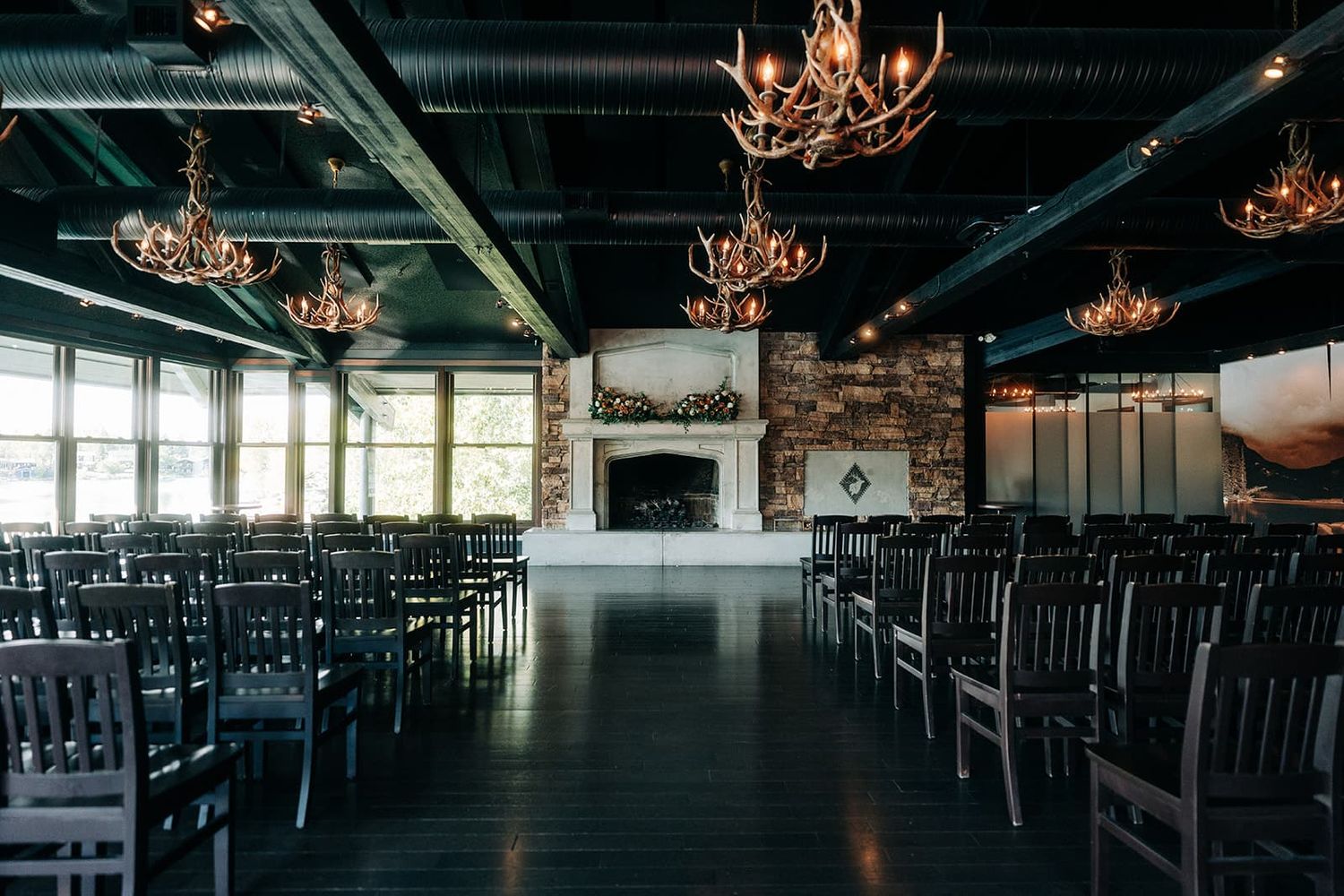 Elegant wedding venue with antler chandeliers, rows of dark wood chairs, and a stone fireplace altar.