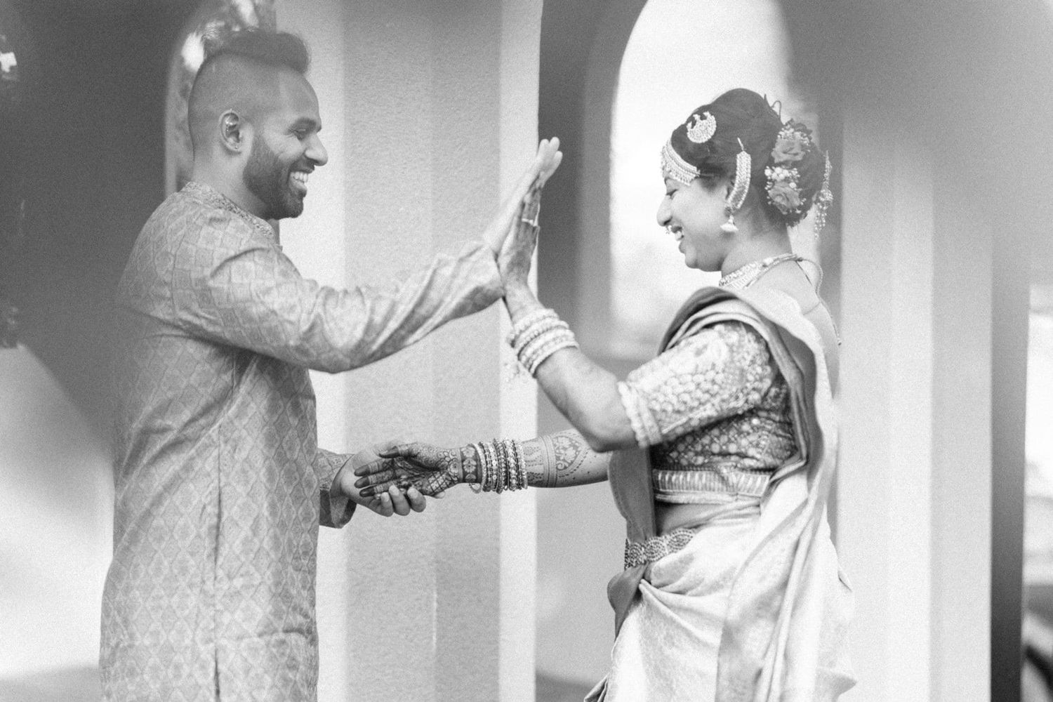 Black and white photo of Indian couple in traditional attire sharing a joyful high-five moment at a wedding ceremony.