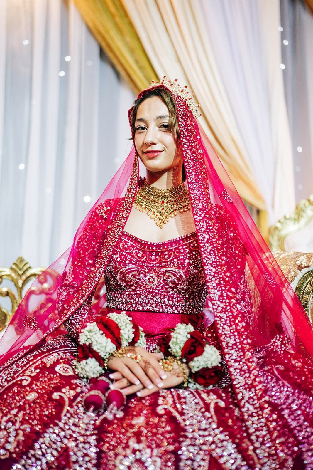 A bride in a red embroidered lehenga with gold jewelry and dupatta sits gracefully during an Indian wedding ceremony.