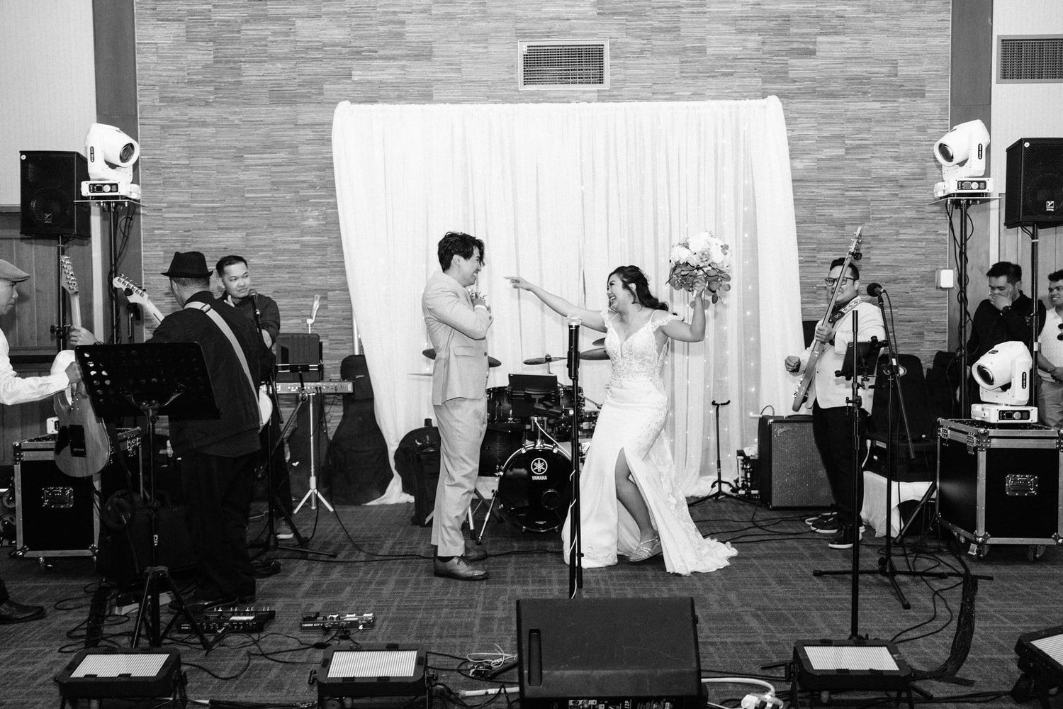 Bride and groom share first dance at wedding reception with live band performing on stage in black and white photo.