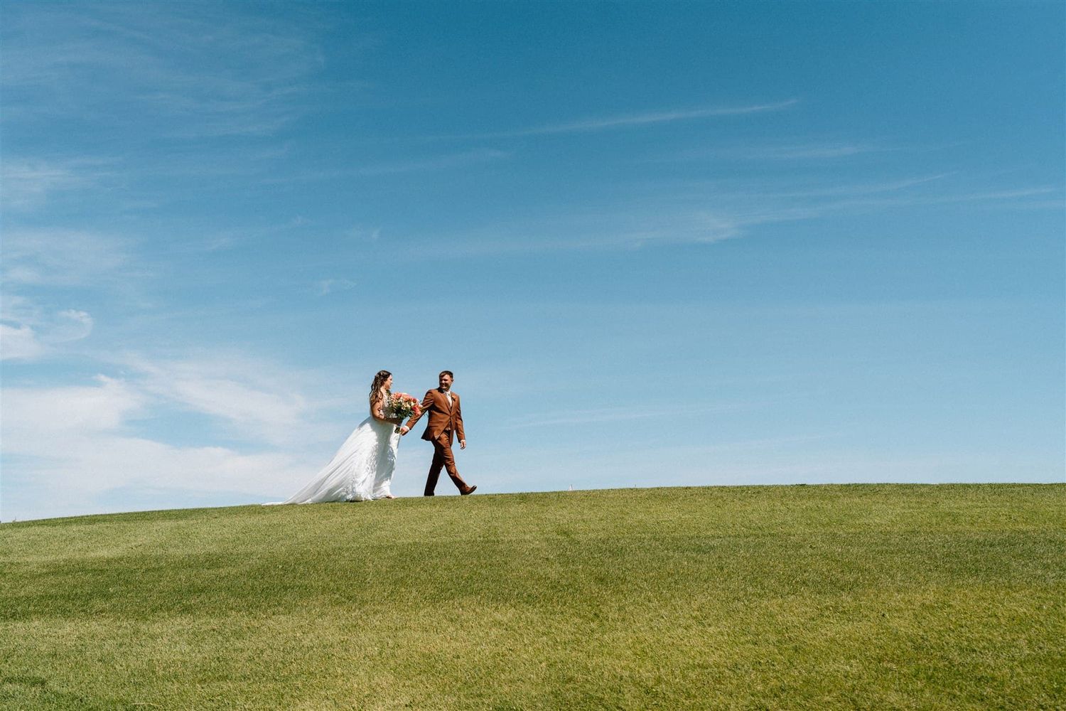 Bride and groom walk hand in hand across a lush green hill under a bright blue sky on their wedding day.