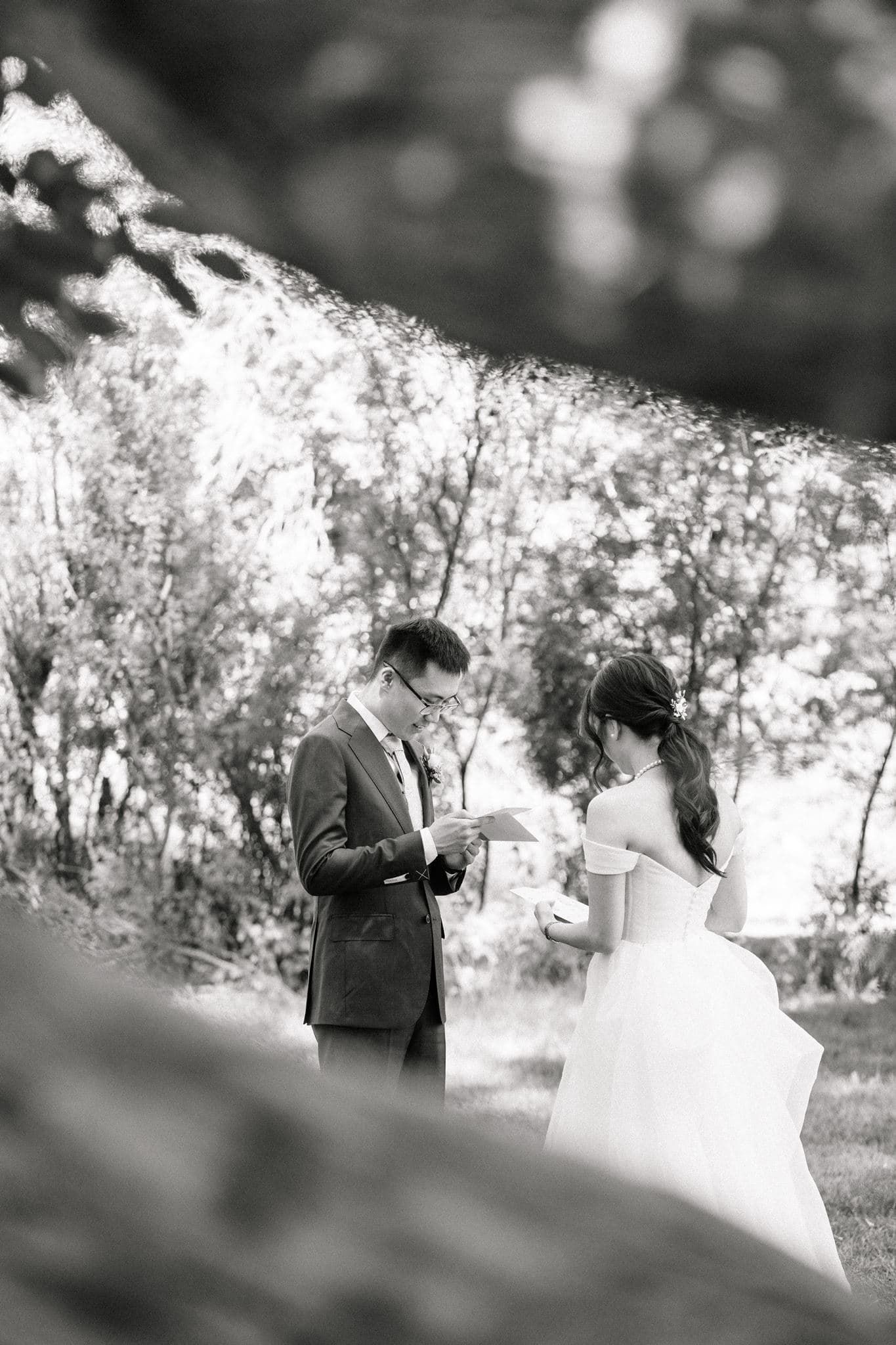 Black and white photo of a bride and groom exchanging wedding vows outdoors among lush green trees.