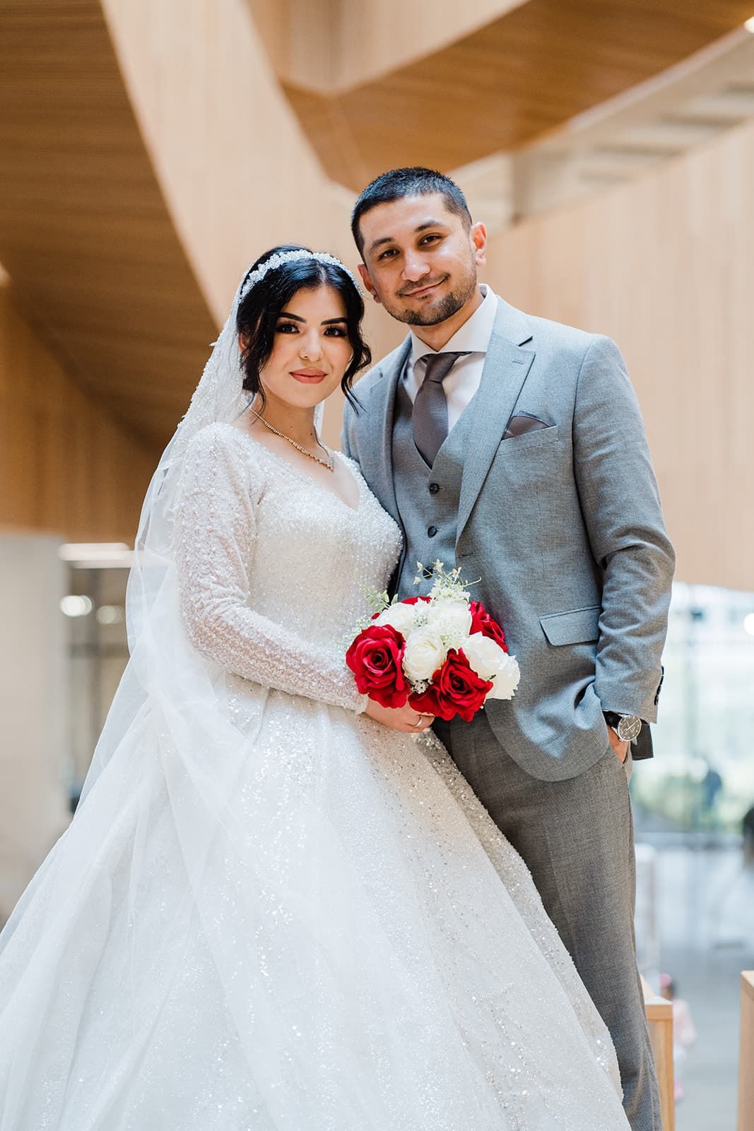 Bride and groom posing together, she in white beaded gown holding red and white bouquet, he in gray suit.