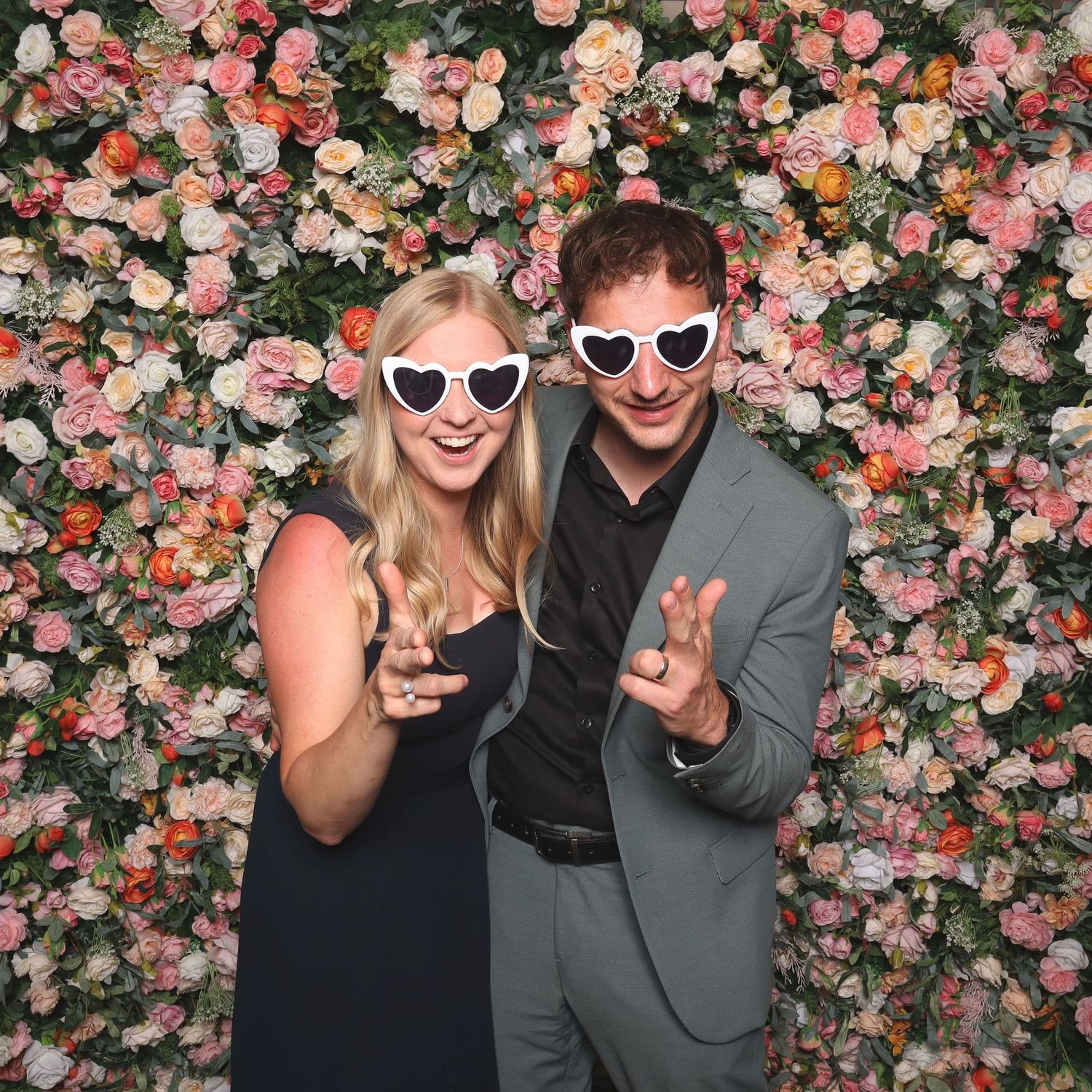 Couple wearing heart-shaped sunglasses posing in front of a floral wall backdrop.