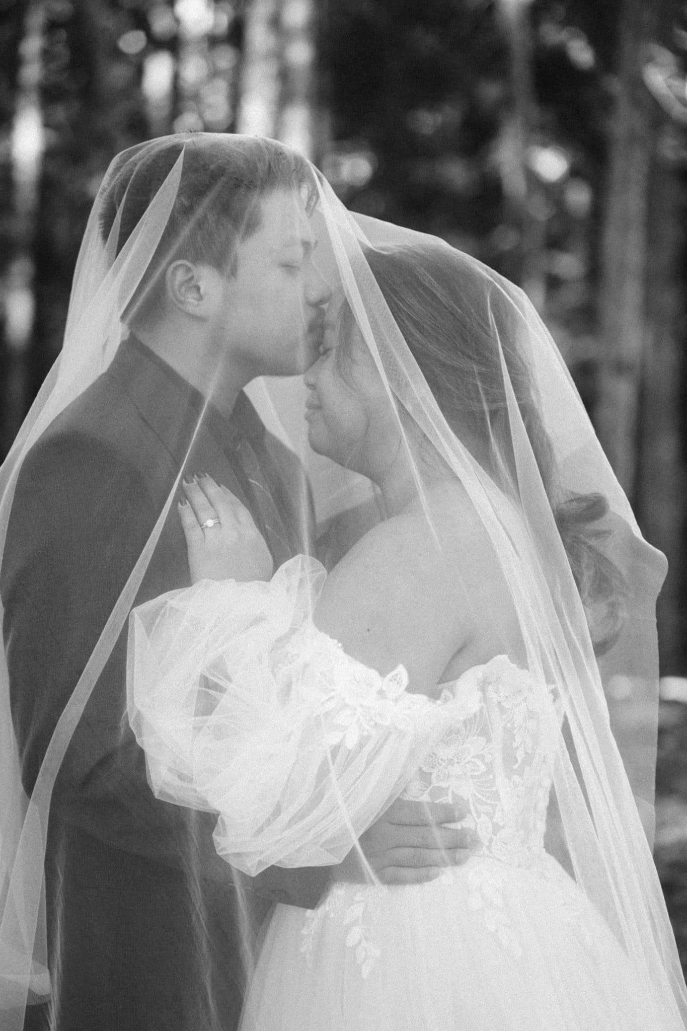 Black and white photo of a bride and groom sharing a romantic kiss under a flowing wedding veil outdoors.