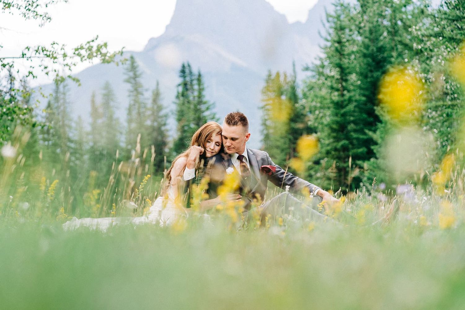 Couple sitting together in a lush green meadow with wildflowers and mountain peaks in the background.