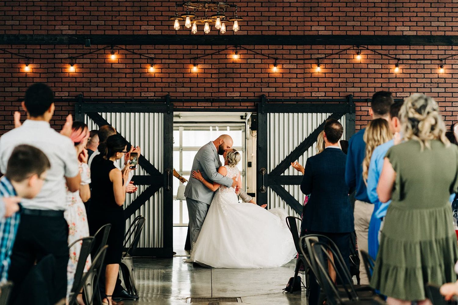 Bride and groom share a kiss at wedding reception in rustic brick venue with string lights and barn doors.