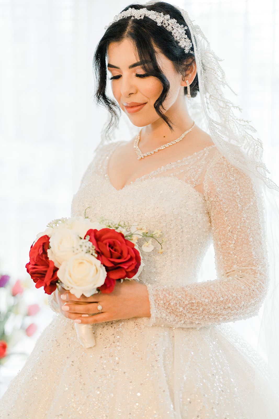 Elegant bride in a sparkly white ball gown holding red and white roses bouquet, wearing a crystal tiara and veil.