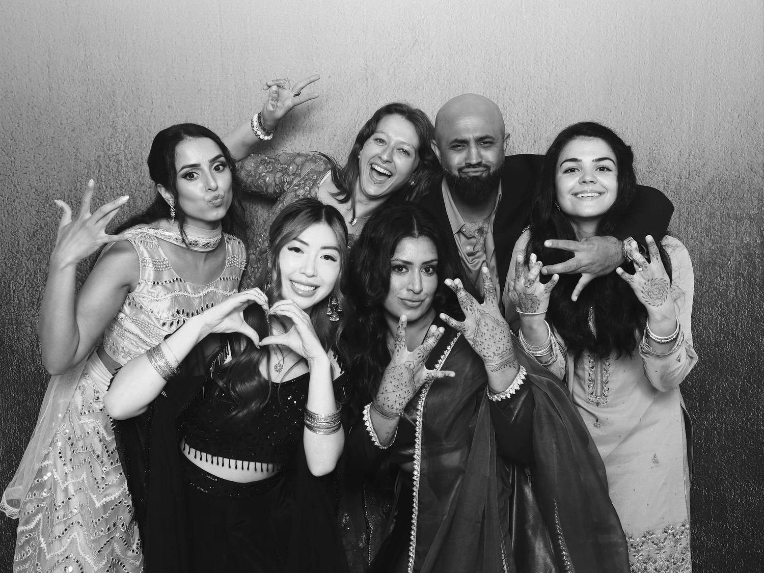 Group of six friends pose playfully making heart signs at a wedding photo booth in black and white.