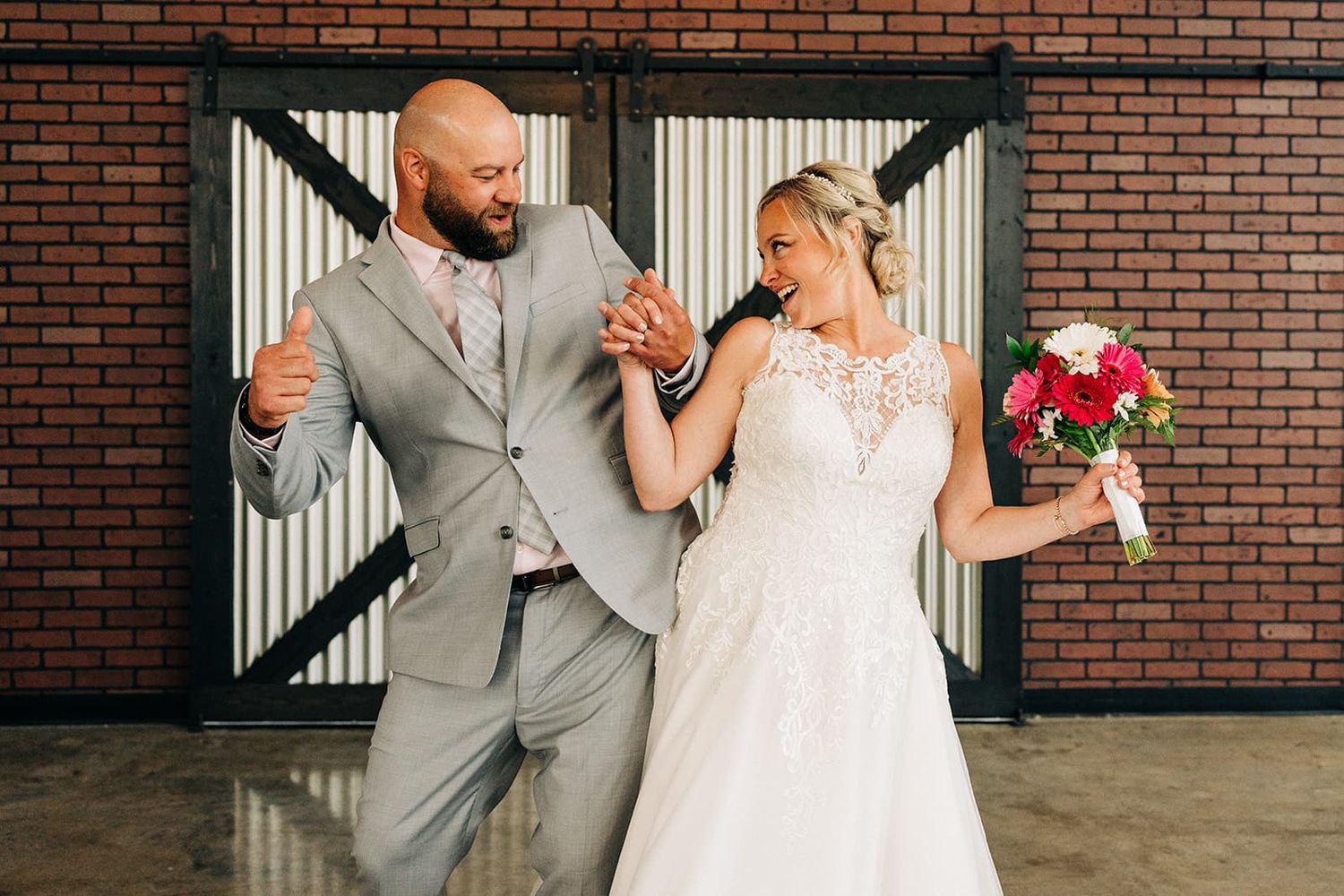 Bride and groom in gray suit dancing joyfully in front of brick wall with pink bouquet.