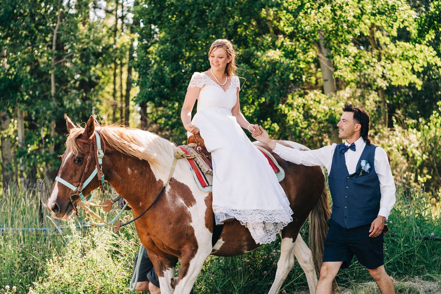 Bride in white wedding dress riding a brown and white horse, guided by groom in vest and bow tie outdoors.