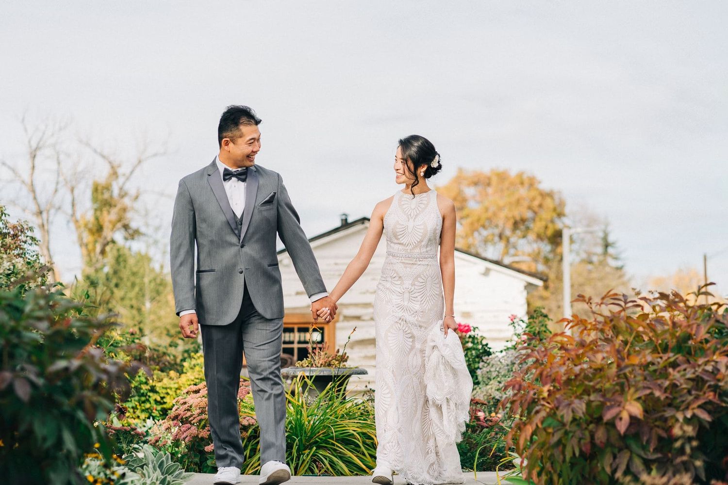 Bride and groom holding hands walking through a lush garden, both smiling in elegant wedding attire.