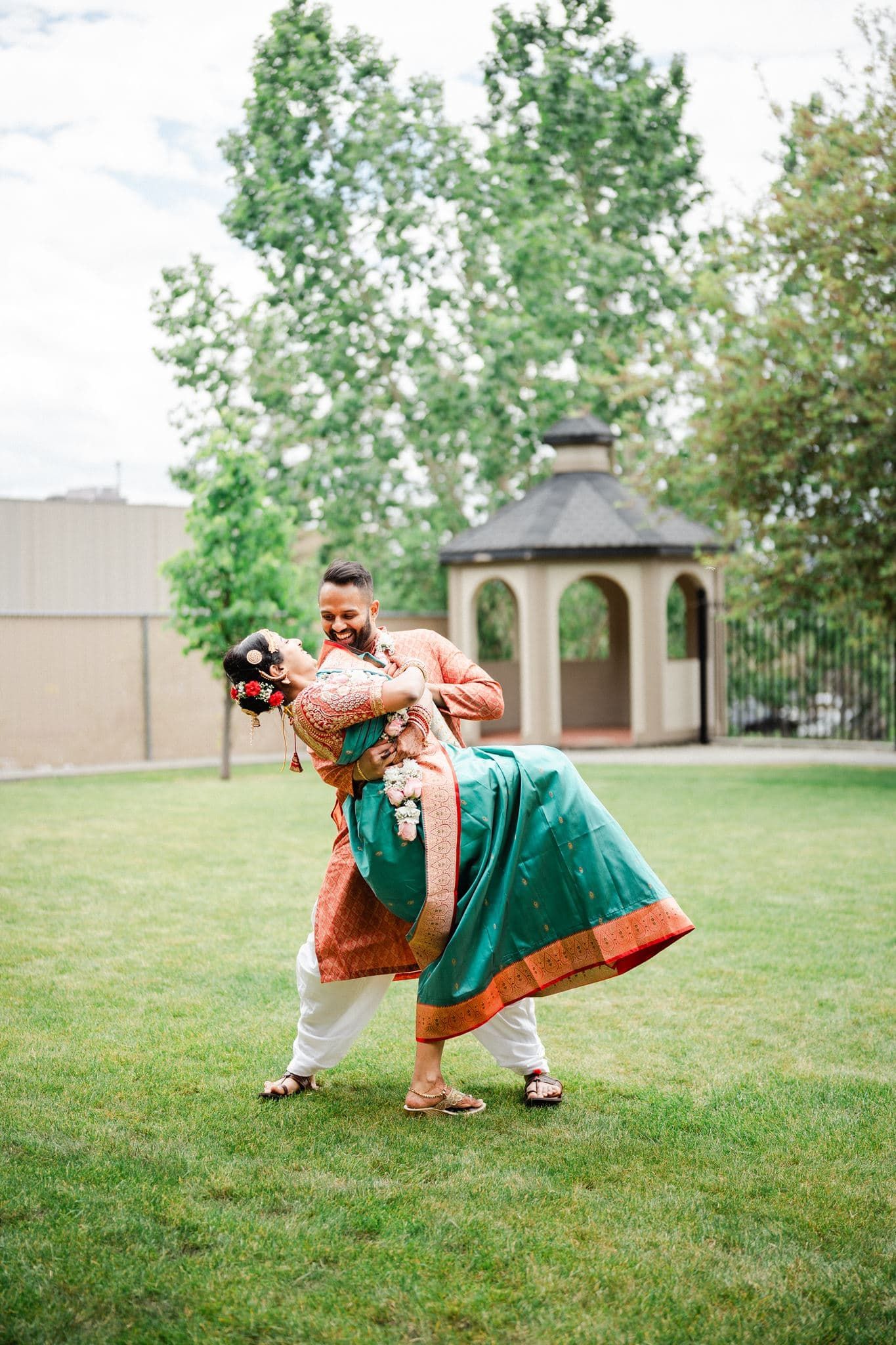 Couple in traditional Indian wedding attire dancing outdoors on green lawn with gazebo in background.
