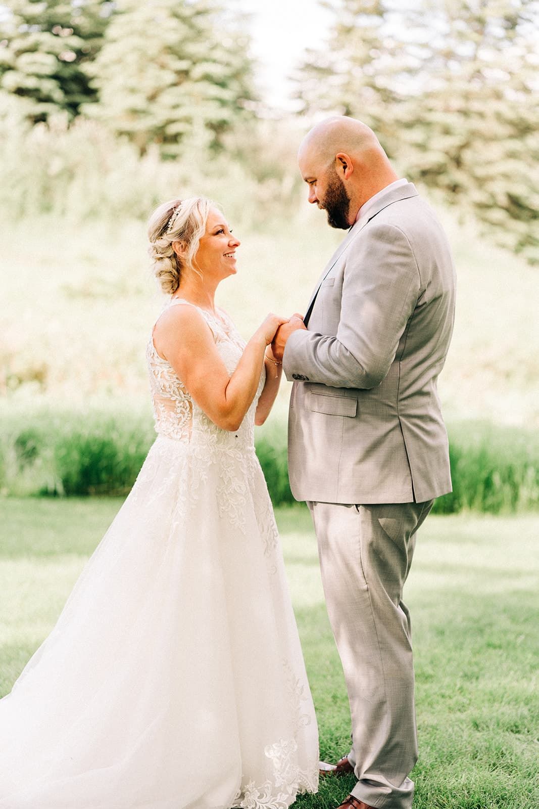 Bride in lace gown and groom in gray suit hold hands and share a joyful moment outdoors on their wedding day.
